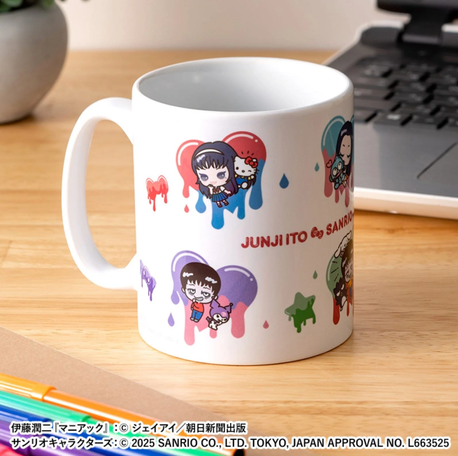 White mug printed with multiple chibi Junji Ito × Sanrio characters and colorful drip shapes, photographed on a wooden desk near a laptop.