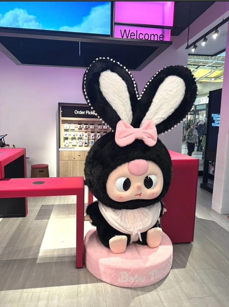 Large plush-like Baby Three bunny statue with tall black-and-white ears and a pink bow, displayed inside a T-Mobile store with magenta lighting.