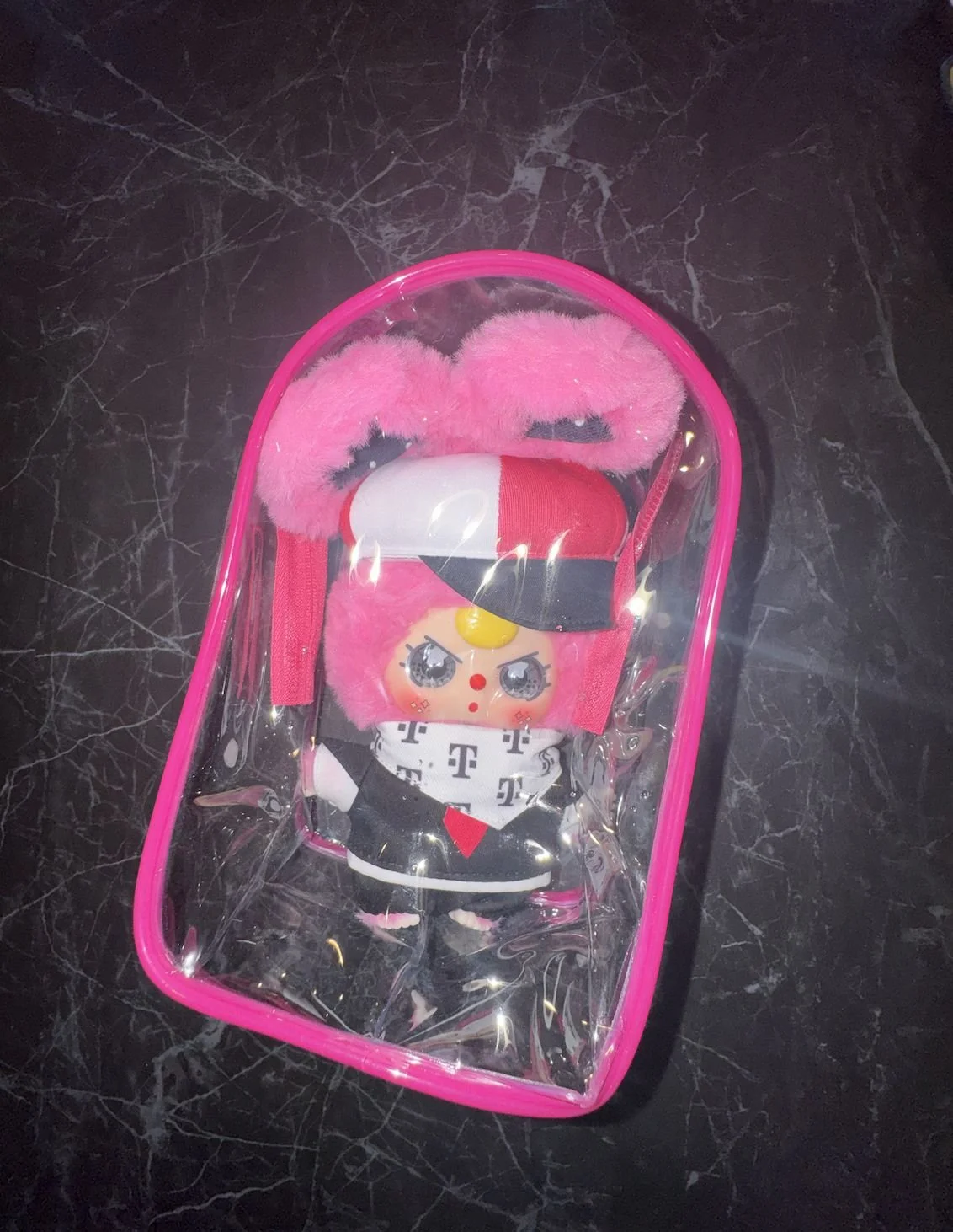 Baby Three Mofi plush inside a clear plastic carrier backpack with bright pink trim on a dark surface.