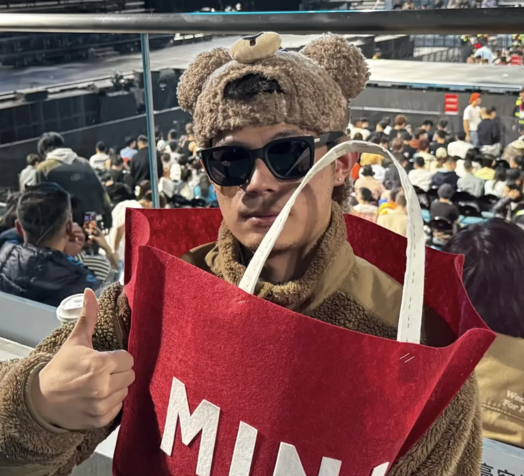 Person at a stadium concert wearing a fuzzy brown bear hat, sunglasses, and a teddy-textured jacket, giving a thumbs-up while sitting in the stands with the stage and crowd behind them; they’re wrapped inside an oversized red felt MINISO shopping bag