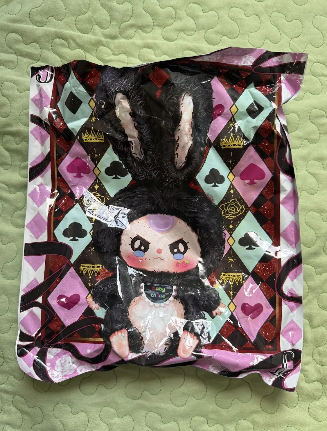 Crinkled retail pouch lying on a green quilt, decorated with harlequin diamonds in mint, pink and black; centre illustration shows Baby Three wearing a fluffy black bunny costume with long ears.