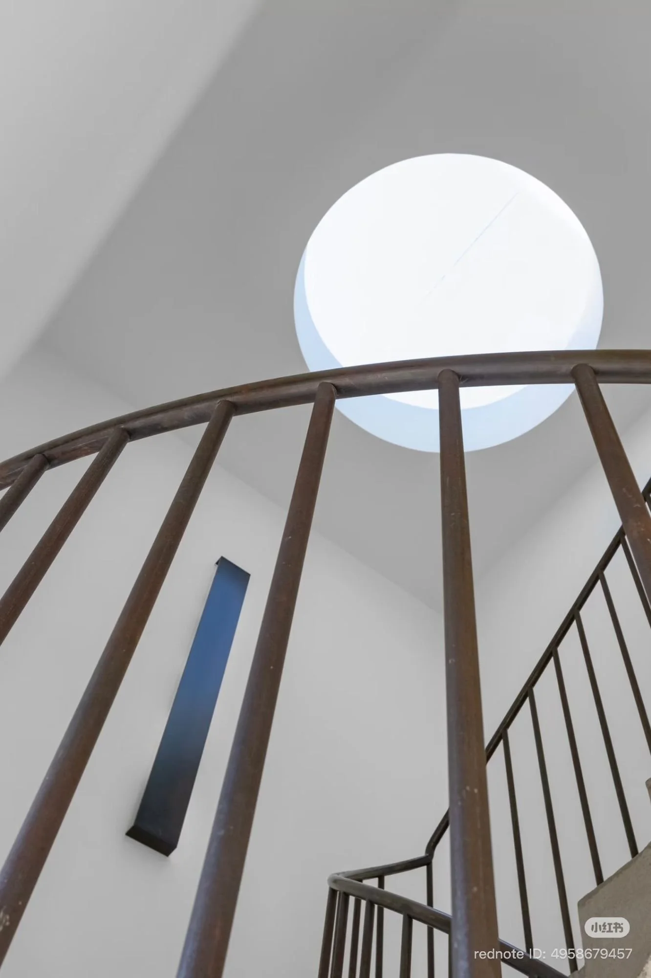 Upward view past a curved metal stair railing toward a circular skylight opening in a white ceiling; the stair continues to the right.