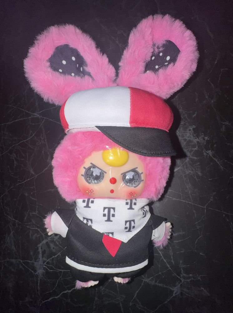 Baby Three Mofi plush with pink fur wearing a red, white, and black cap and a white bandana printed with T logos.