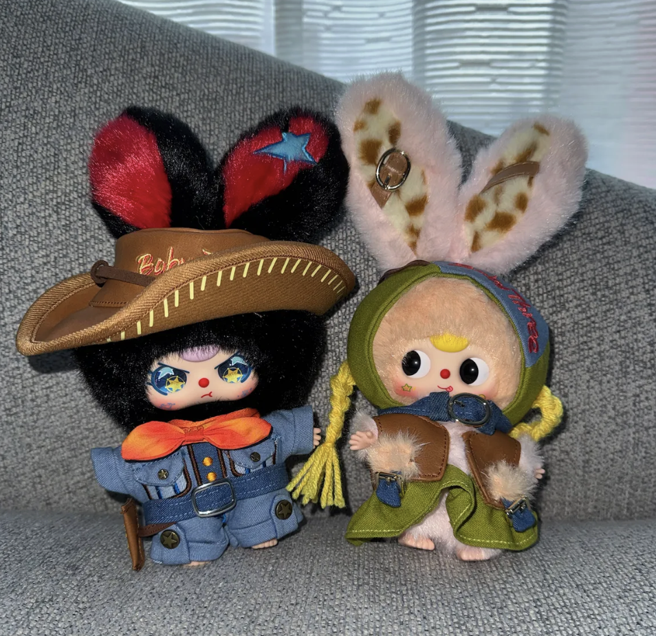 Two Baby Three collectible dolls — one wearing a brown cowboy-style hat and a denim jacket with orange bow tie, the other in a green hood and pink furry ears — posed side-by-side on a gray fabric surface.