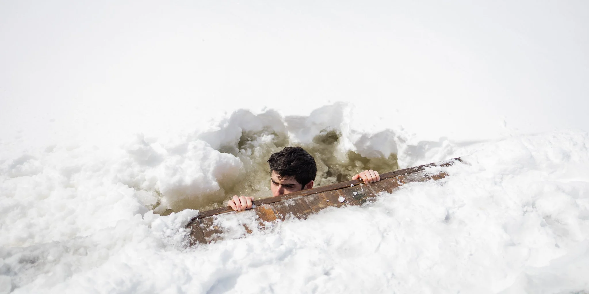 Man climbing out of a hole in the snow while holding onto a wooden edge.