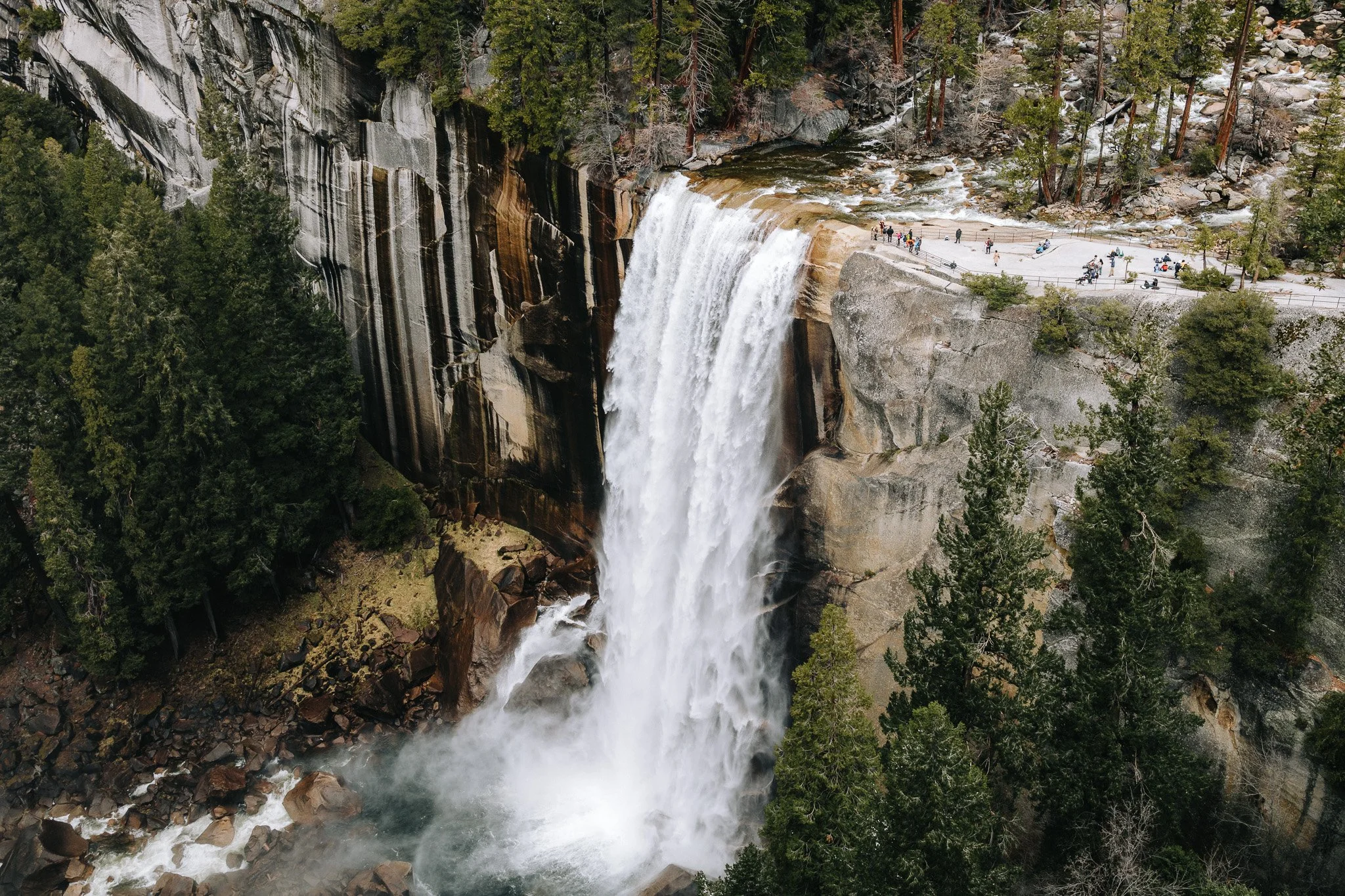 Aerial view of a waterfall flowing over large rocks surrounded by trees, with tourists walking on a viewing platform.