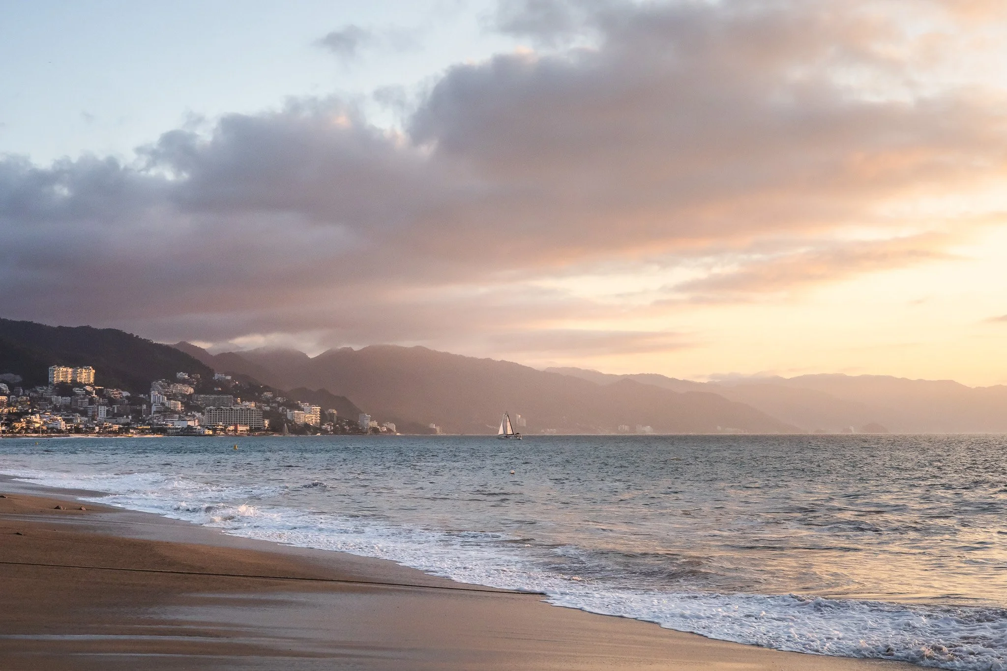 Sunset over a beach with waves, a sailboat on the water, and a city skyline at the foot of mountains