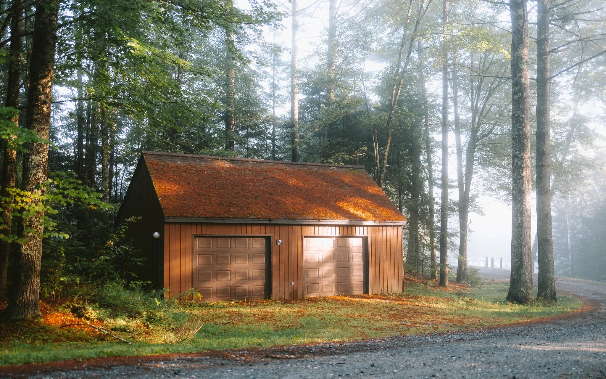 A wooden garage with a rusty, moss-covered roof, situated in a forest clearing with tall trees and sunlight filtering through the foliage.