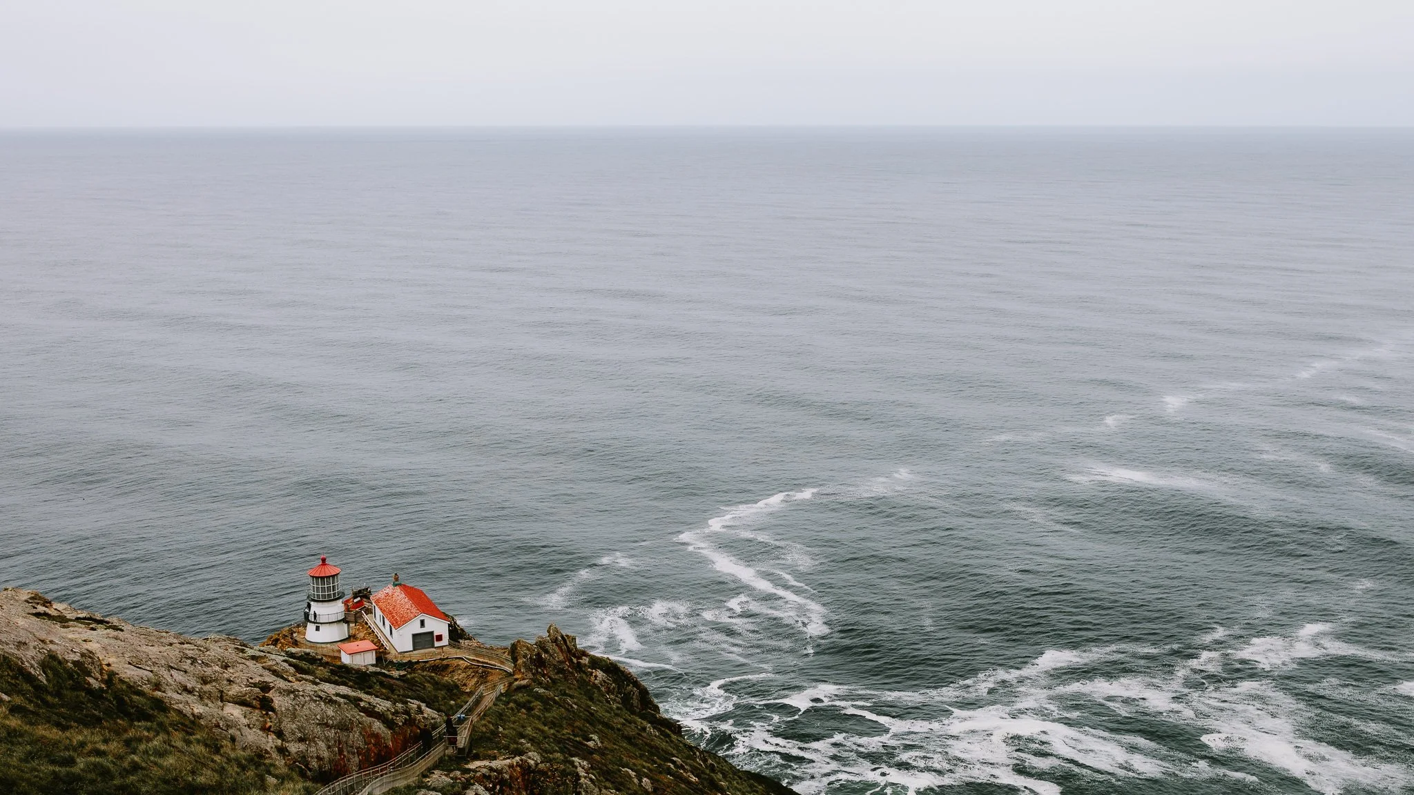 A lighthouse and small building with red roofs on a rocky cliff overlooking the ocean, with waves crashing below.