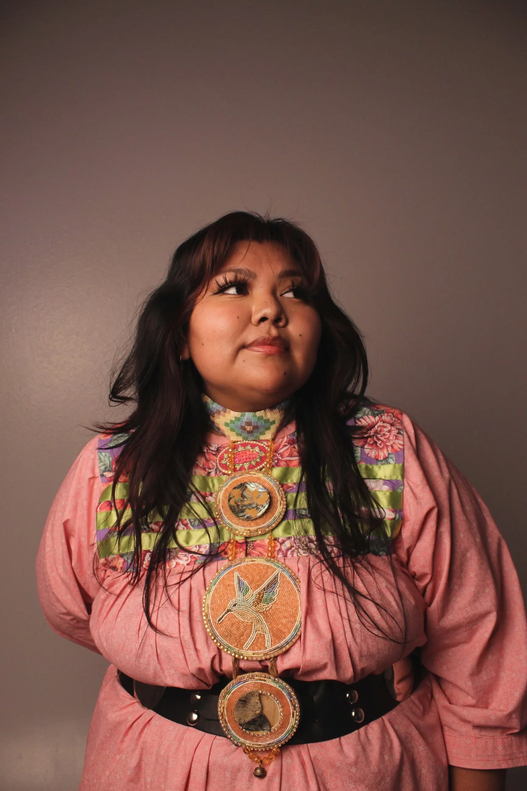 A woman with long dark hair wearing a pink dress and elaborate beaded necklaces with various animal, bird, and nature designs, standing against a neutral background.