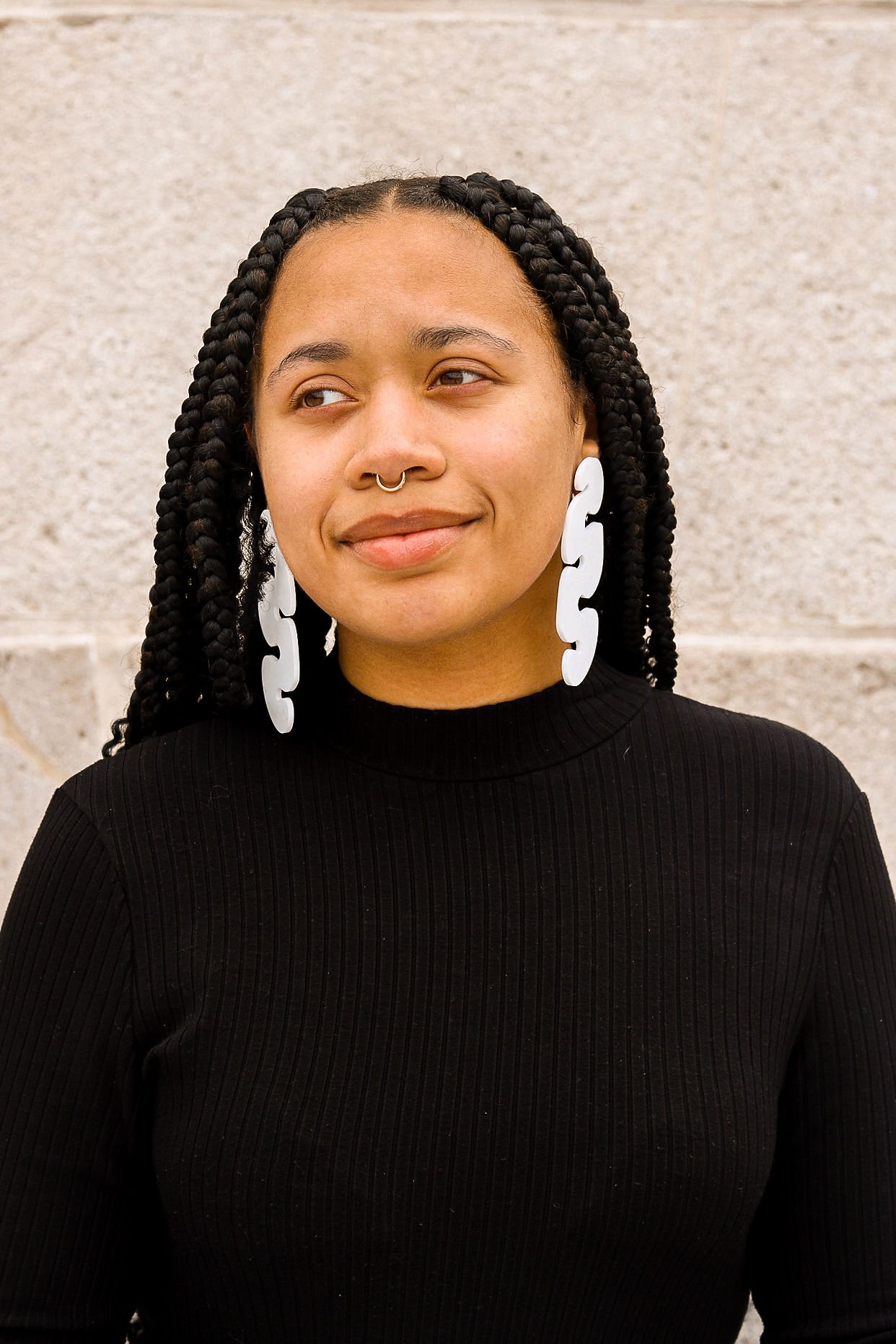 A woman with dark braided hair, white statement earrings, and a septum piercing, wearing a black ribbed turtleneck sweater, standing against a stone wall.