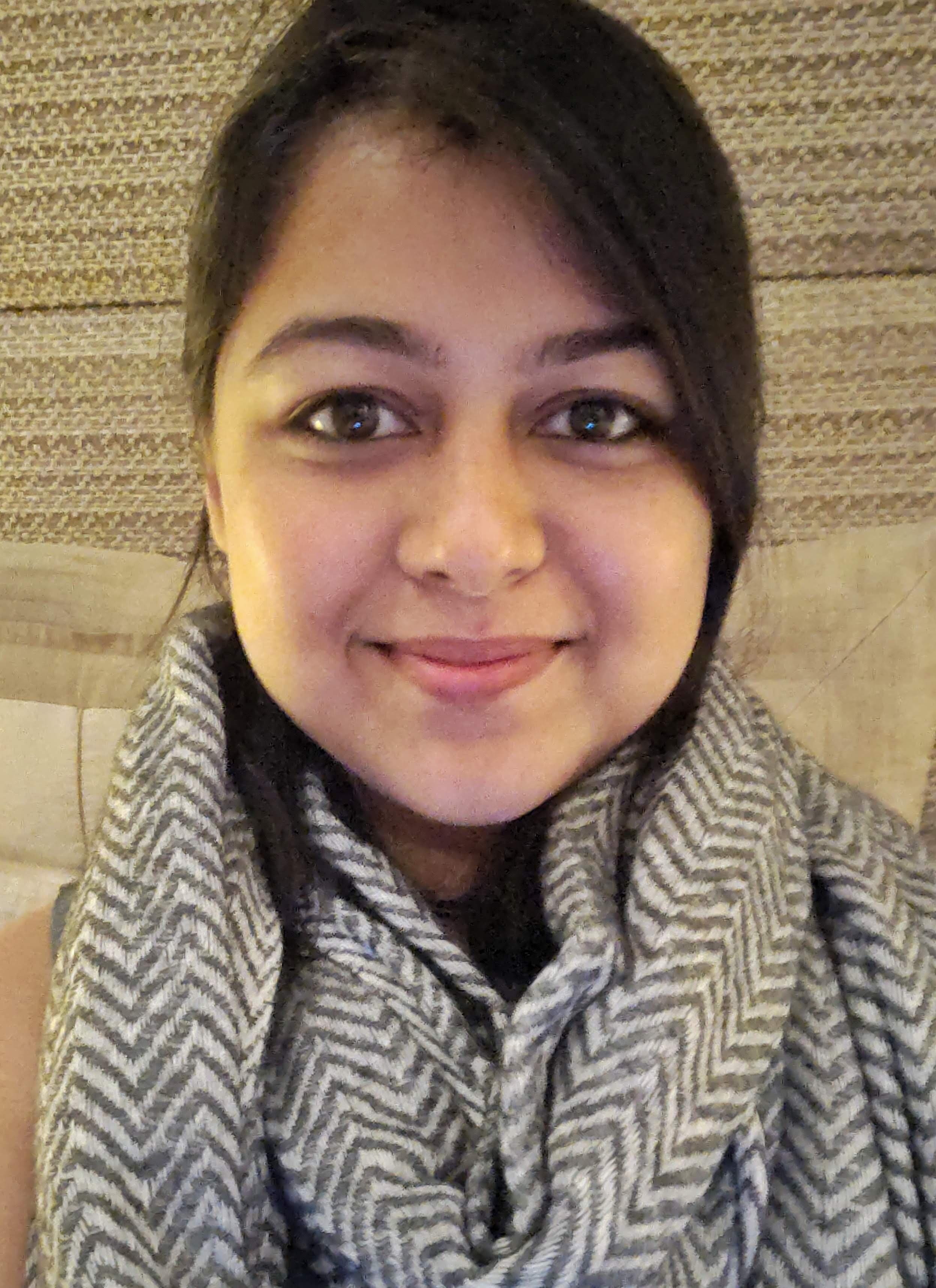 A woman smiling, wearing a striped scarf, with short dark hair, in front of a textured beige wall.