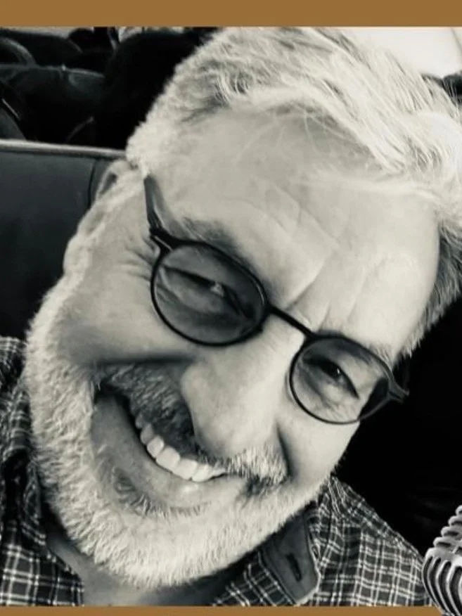 A smiling older man with gray hair, glasses, and a beard, wearing a checkered shirt, in an indoor setting.