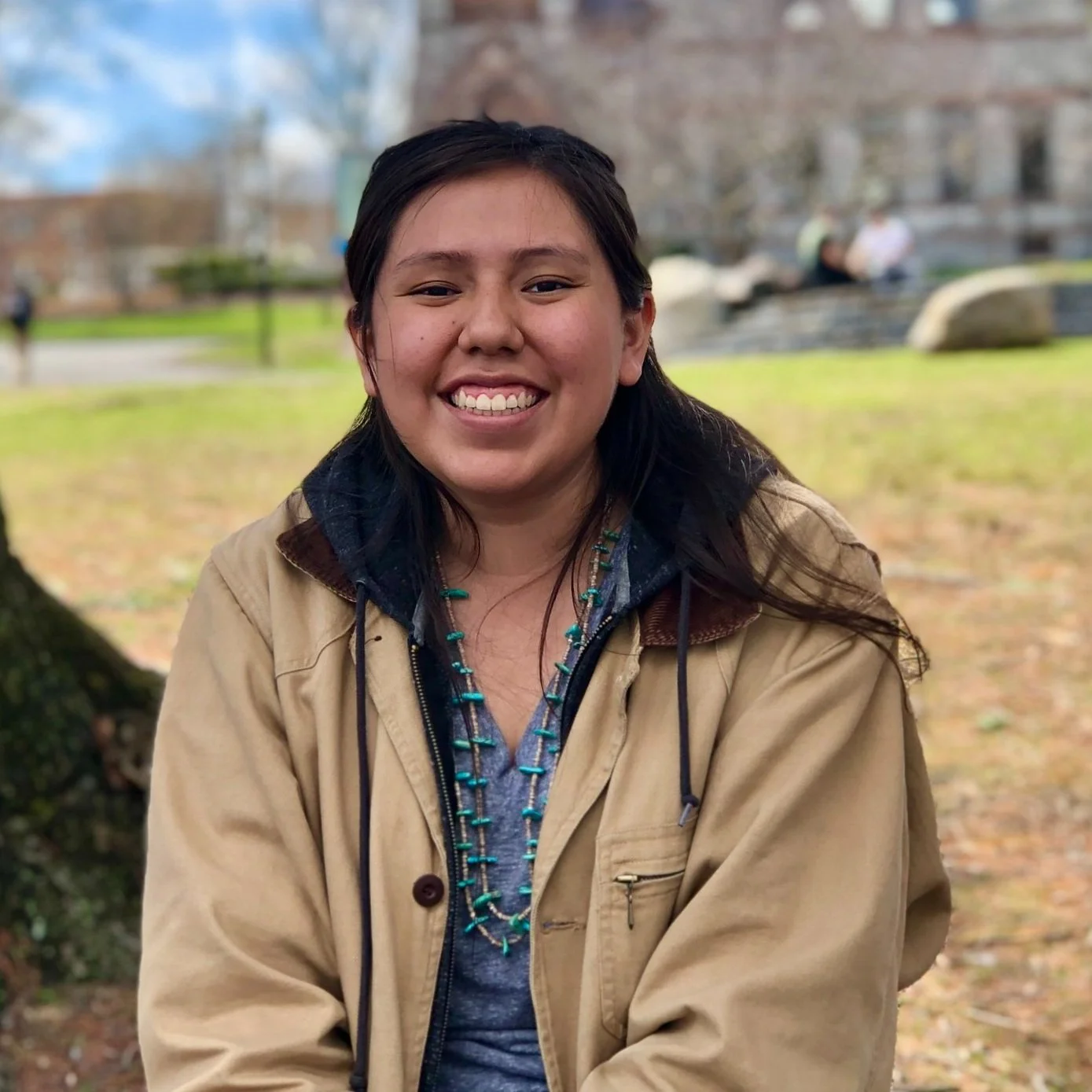 Smiling woman with dark hair and a beaded necklace outdoors in a park during fall.