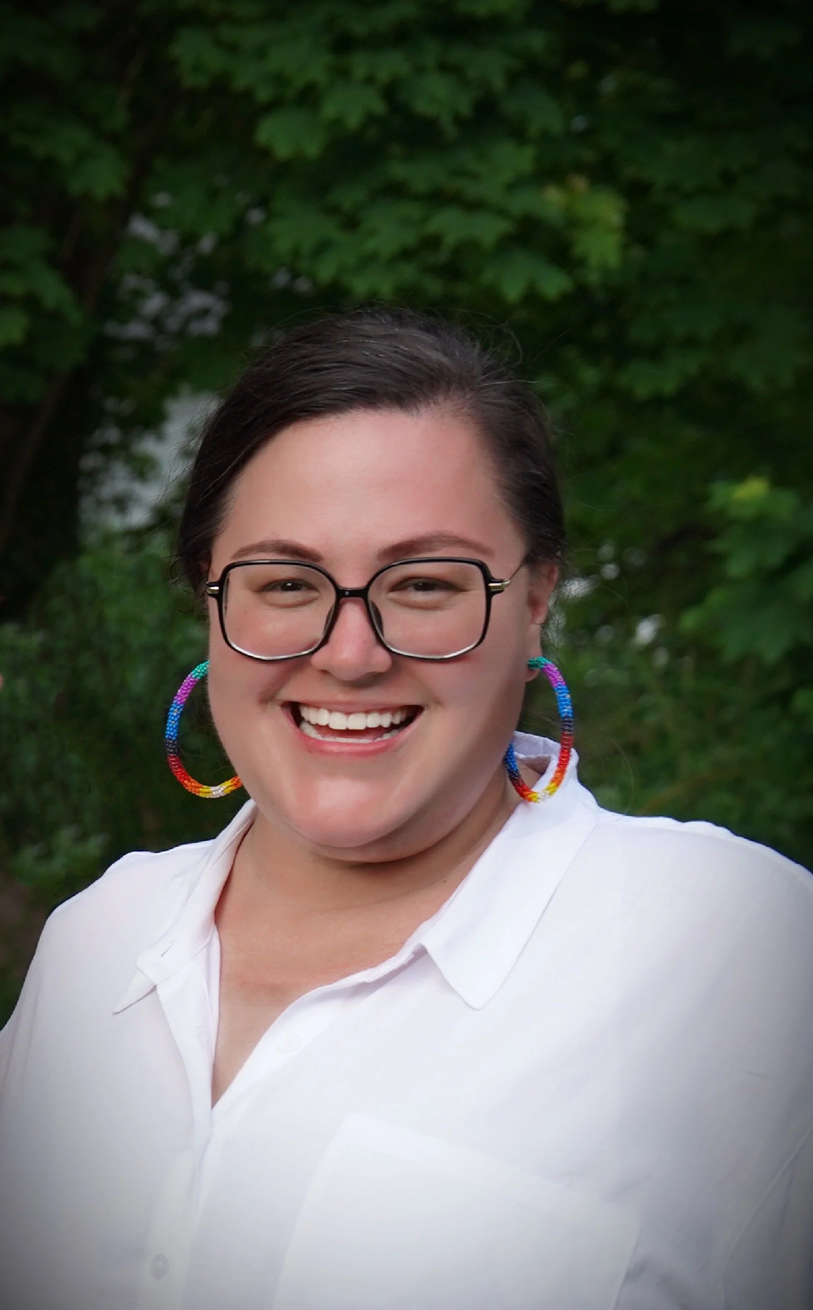 A woman smiling outdoors, wearing glasses, colorful hoop earrings, and a white shirt with a background of green trees.