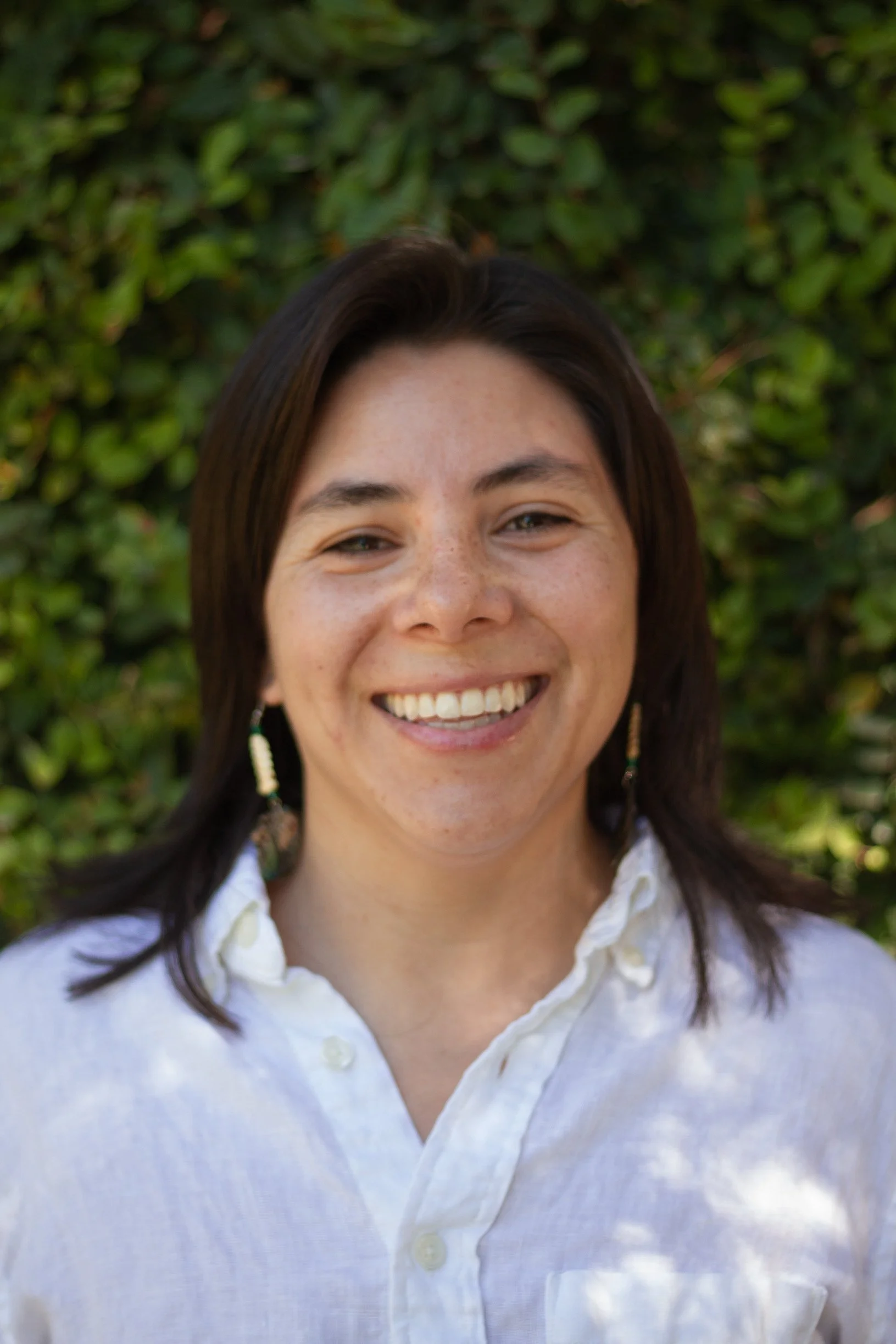 A woman with dark brown hair smiling outdoors in front of a green leafy background, wearing earrings and a white button-up shirt.