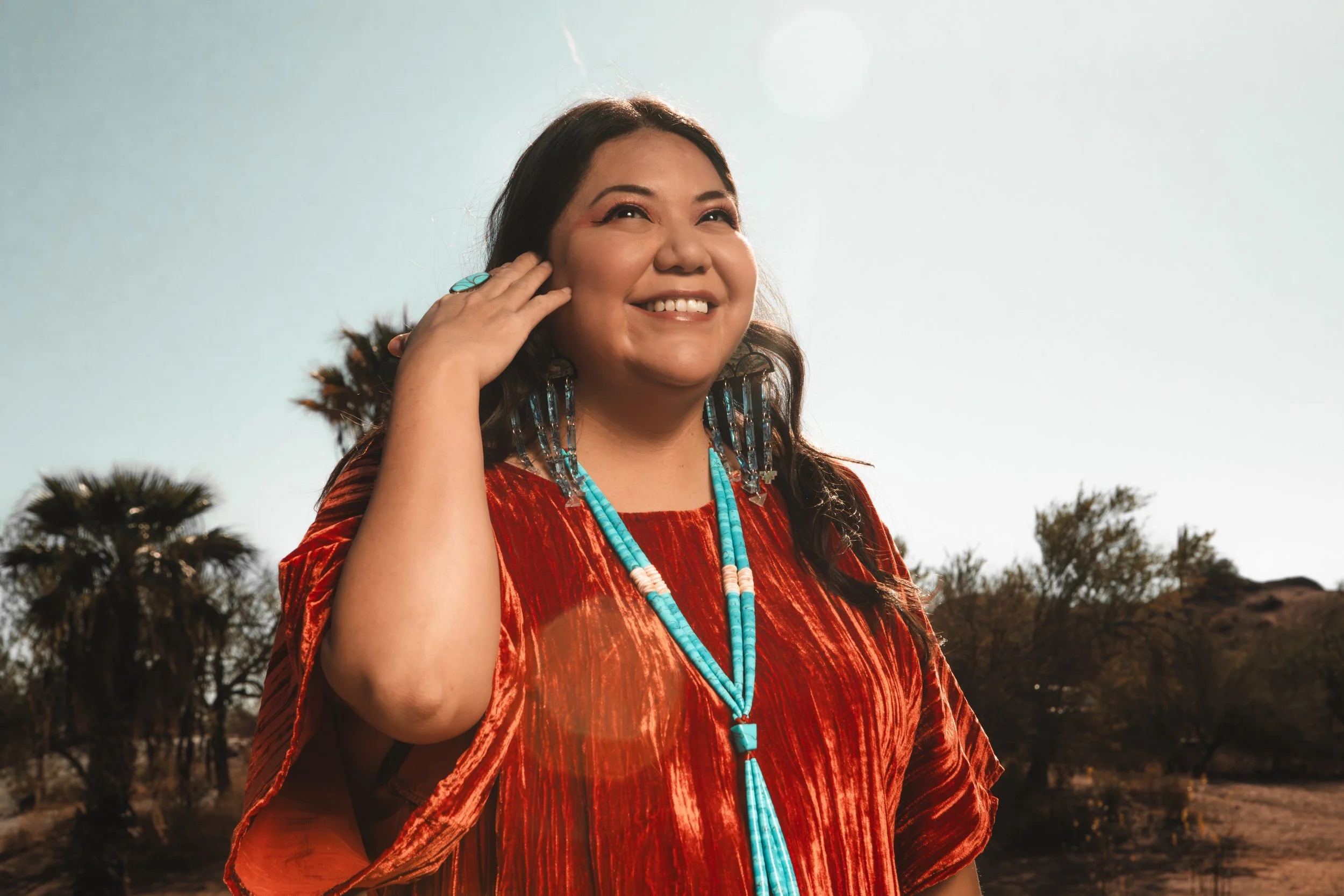 A woman smiling outdoors in a desert landscape with trees, wearing a red dress and turquoise jewelry.