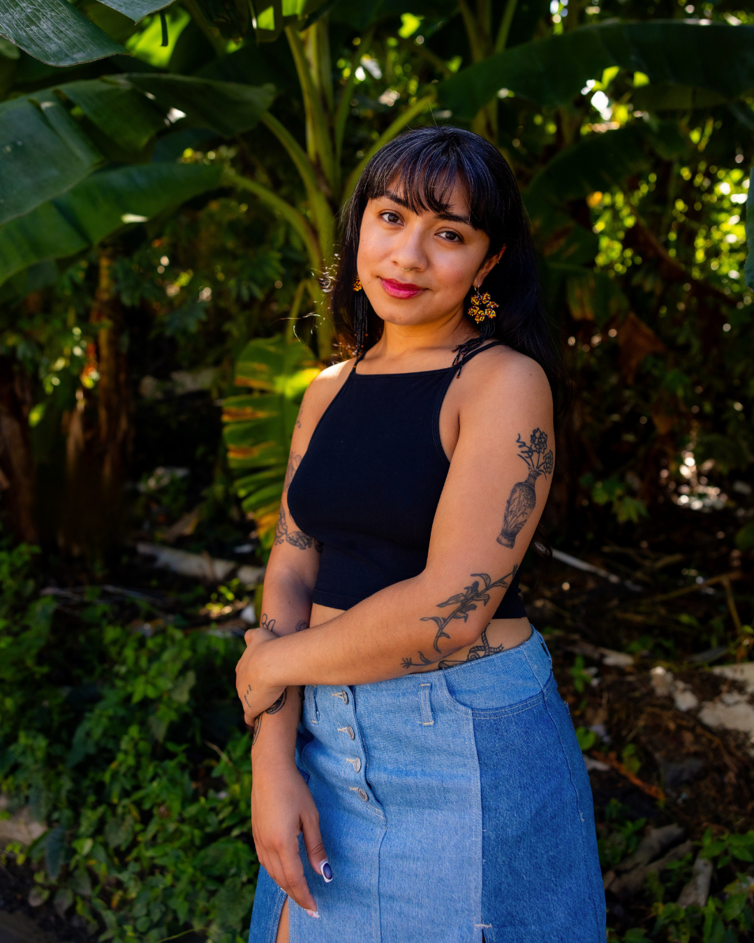 A woman with dark hair and tattoos on her arms wearing a black sleeveless top and a denim skirt, standing outdoors with lush green foliage in the background.
