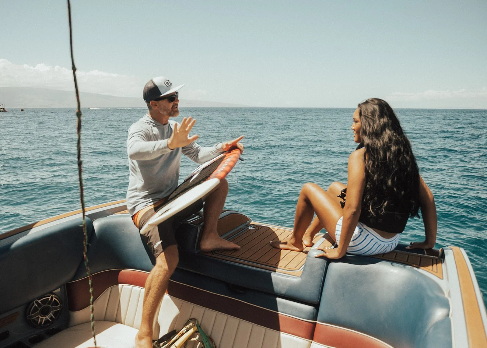 A man and a woman on a boat in the ocean, with the man sitting on the boat's edge and the woman sitting on a padded seat, facing each other, having a conversation.