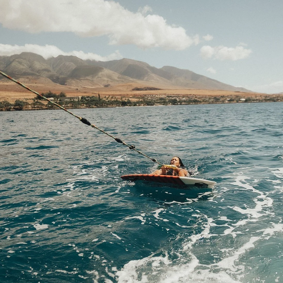 A woman on a surfboard being pulled by a boat in a large body of water with mountains and a partly cloudy sky in the background.