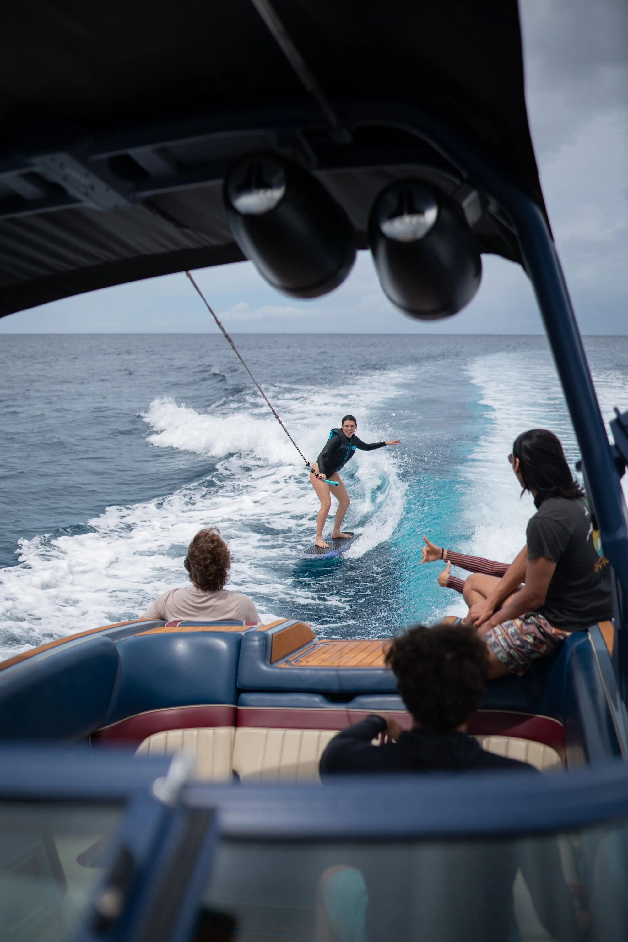 People on a boat engaged in wakeboarding on the water, with one person being pulled on a wakeboard and others watching, under cloudy skies.