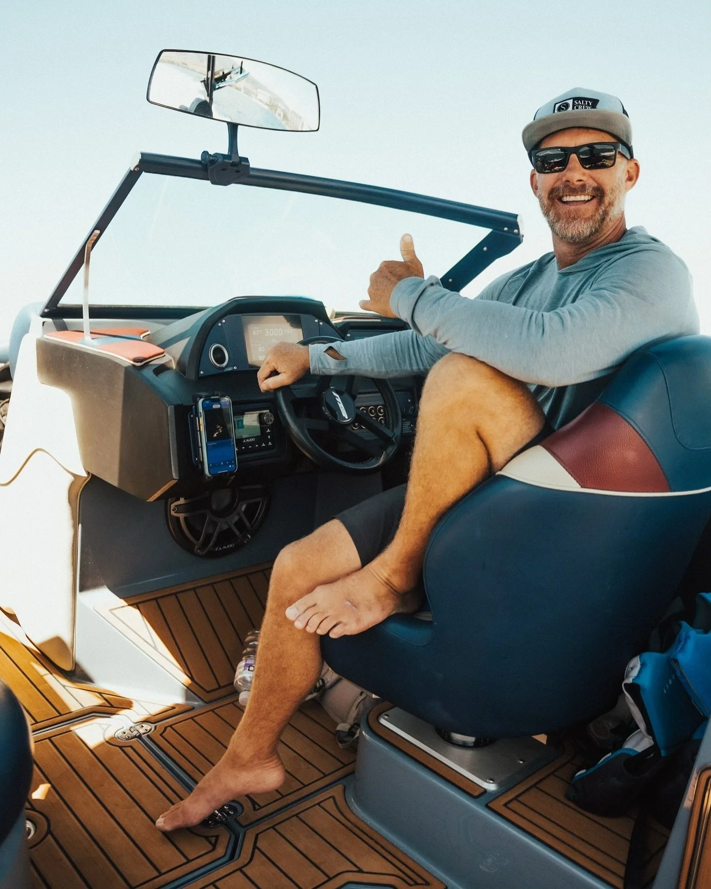 A man sitting in the captain's seat of a boat, smiling, wearing a gray hoodie, sunglasses, and a gray cap. His left foot is resting on the boat's controls, and his right foot is on the floor. The boat's dashboard has a GPS screen and a phone holder. The boat interior features wood-like flooring and a blue leather seat.