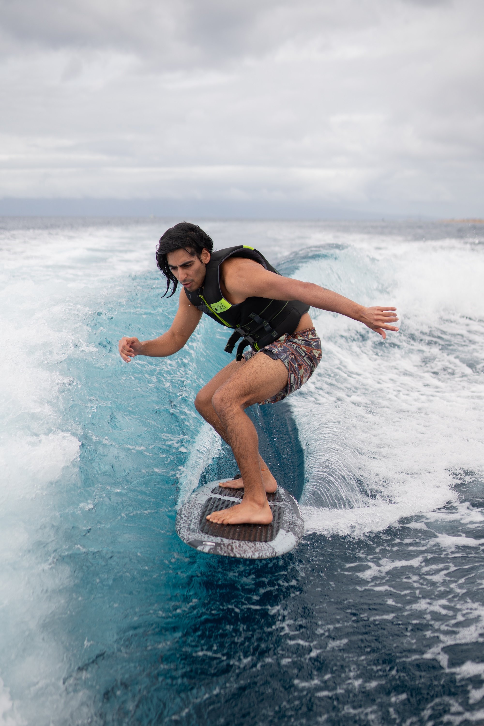 A man surfing on a wave in the ocean, wearing a black life jacket and colorful swim trunks under a cloudy sky.