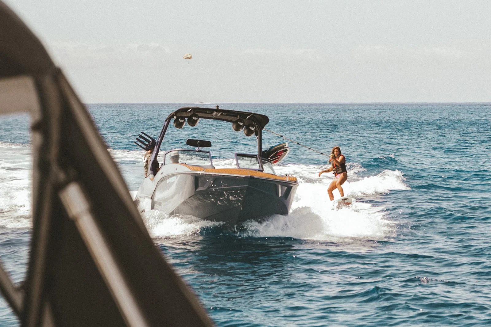 A woman waterskiing behind a motorboat in the ocean on a sunny day, viewed from another boat or structure at the left edge of the image.