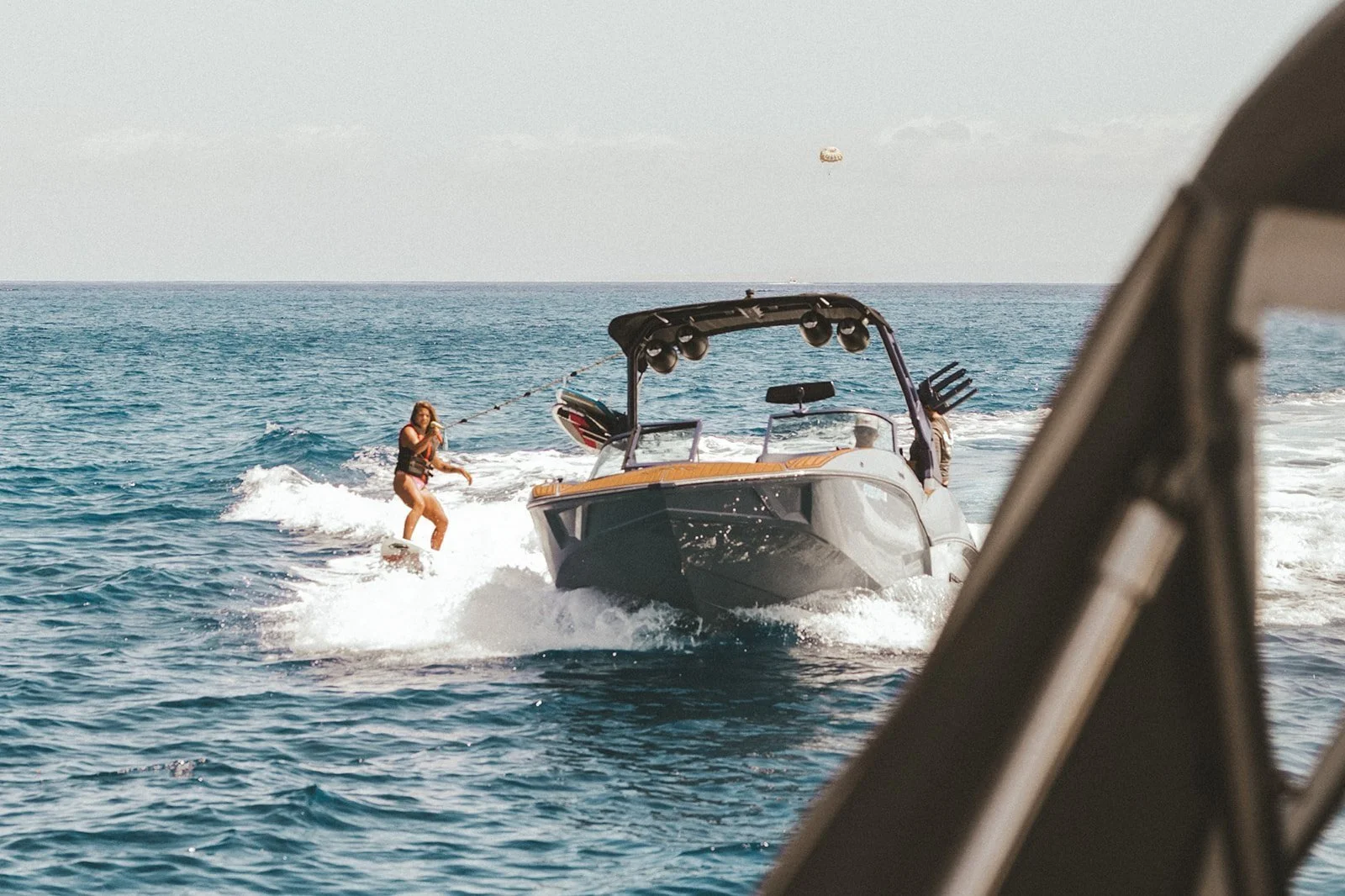 A woman water skis behind a boat on the ocean, with a parasail in the sky in the background.