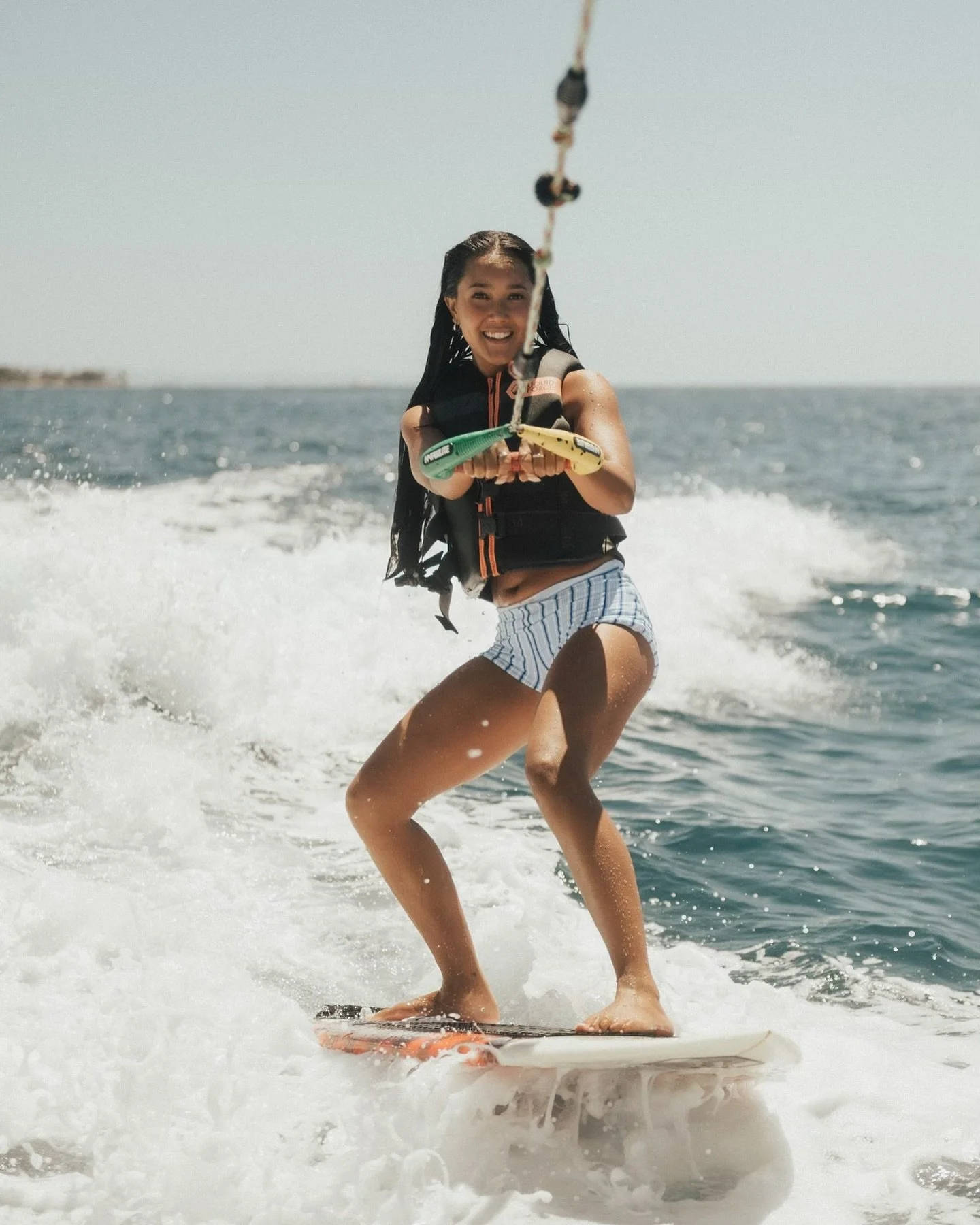 Young woman surfing on a wave in the ocean, wearing a life vest and striped shorts, smiling.
