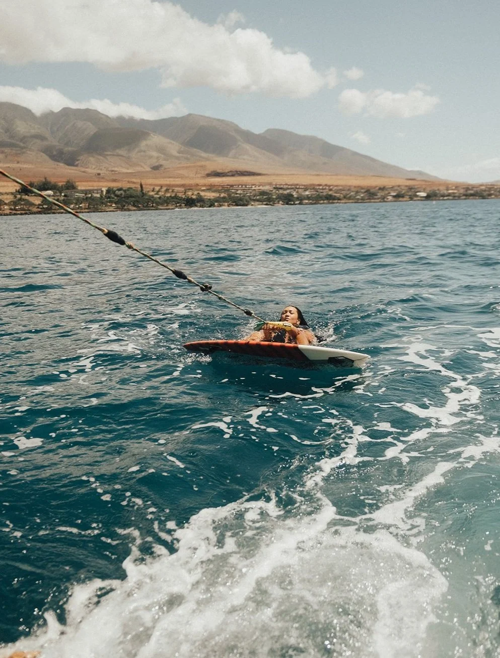 A woman on a surfboard, holding onto a tow rope, being pulled through a body of water with mountains and a cloudy sky in the background.