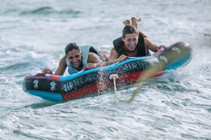 Two women are riding on a blue, red, and black inflatable tube in the water, holding on and smiling.