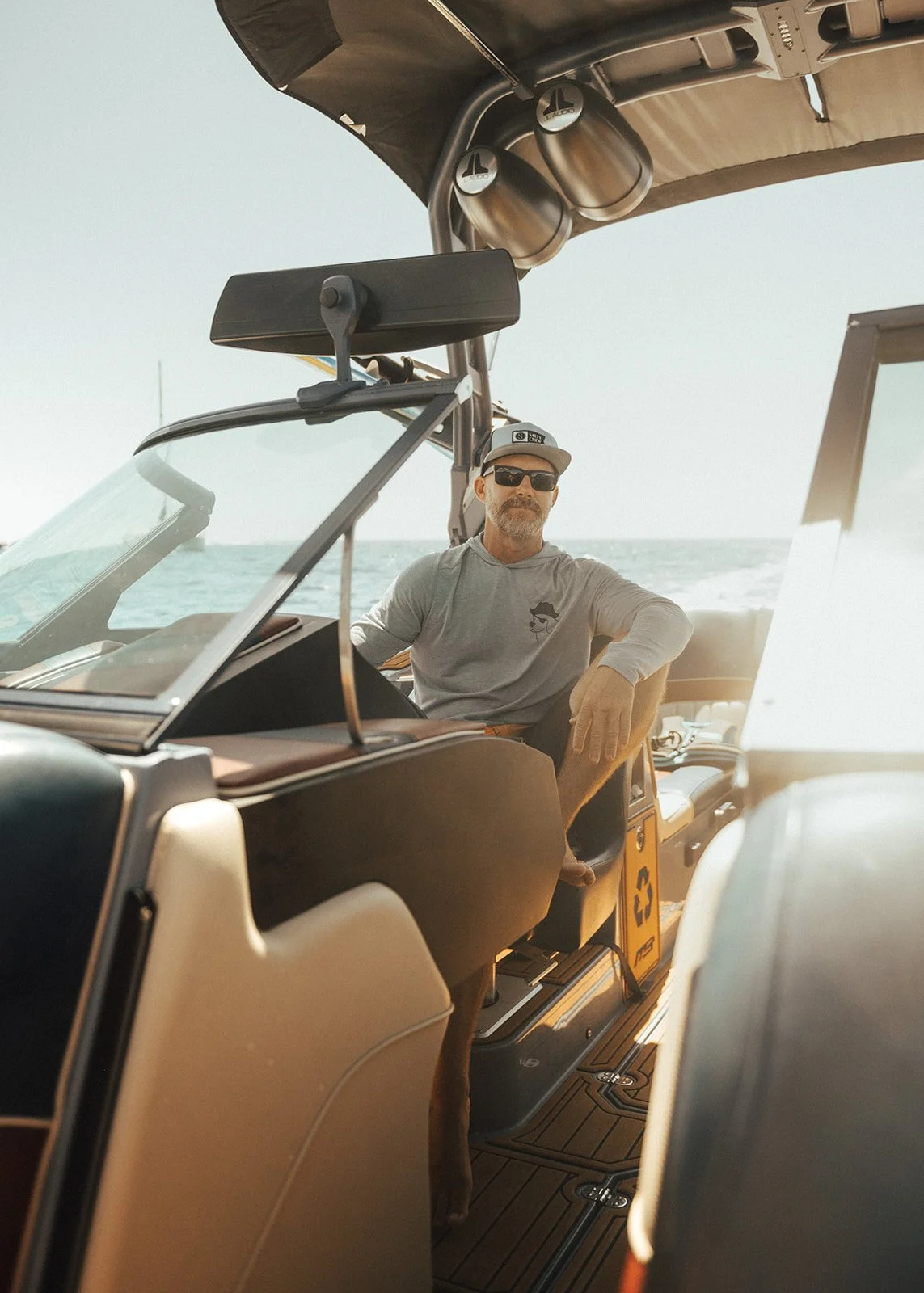 A man sitting on a boat with the ocean in the background, wearing sunglasses, a gray hoodie, and a cap, with boat equipment and a recycling bin nearby.
