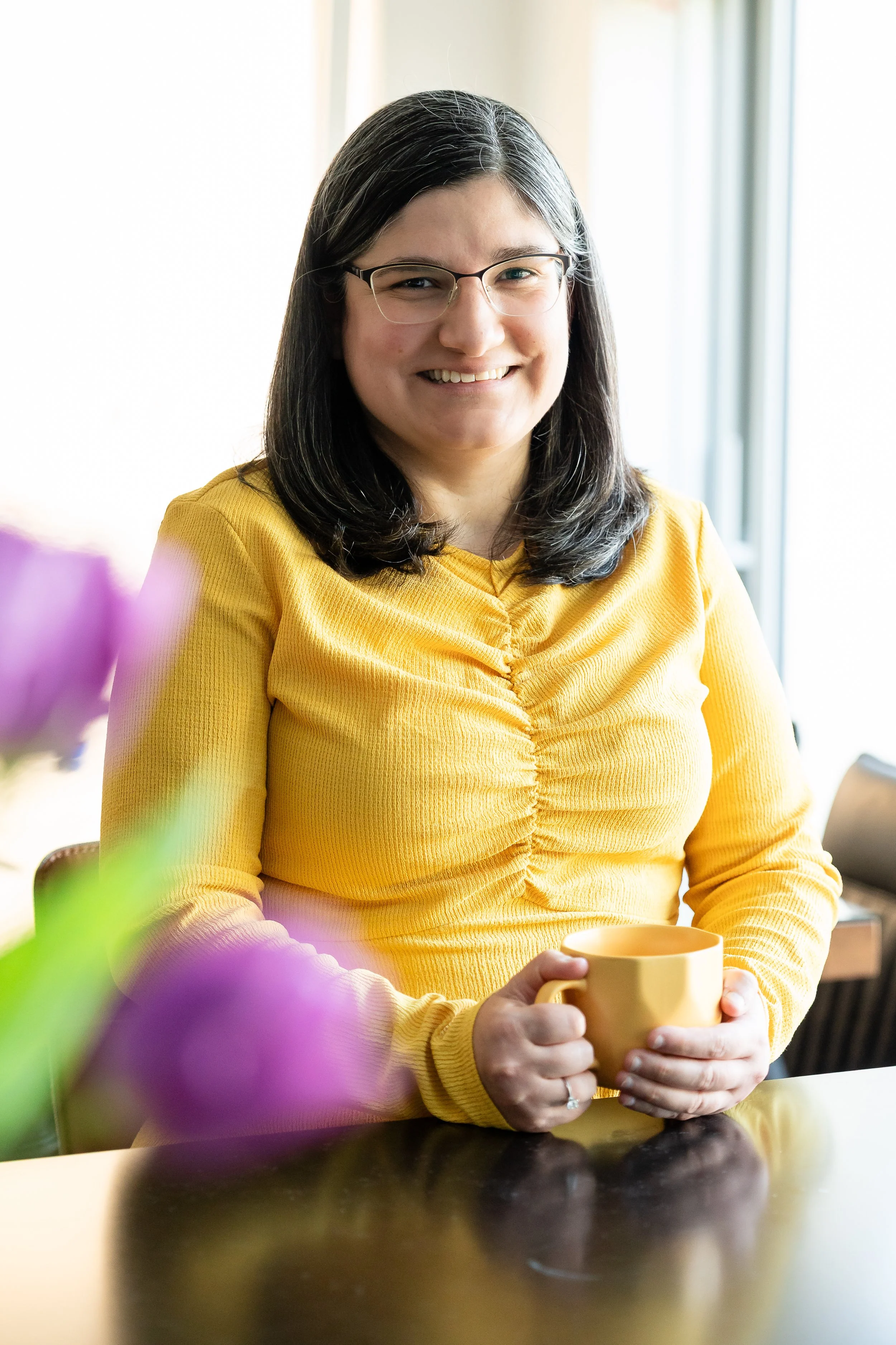 Woman smiling dressed in a yellow dress holding a yellow mug in a kitchen with a clear worktop and a purple tulip popping into the photo