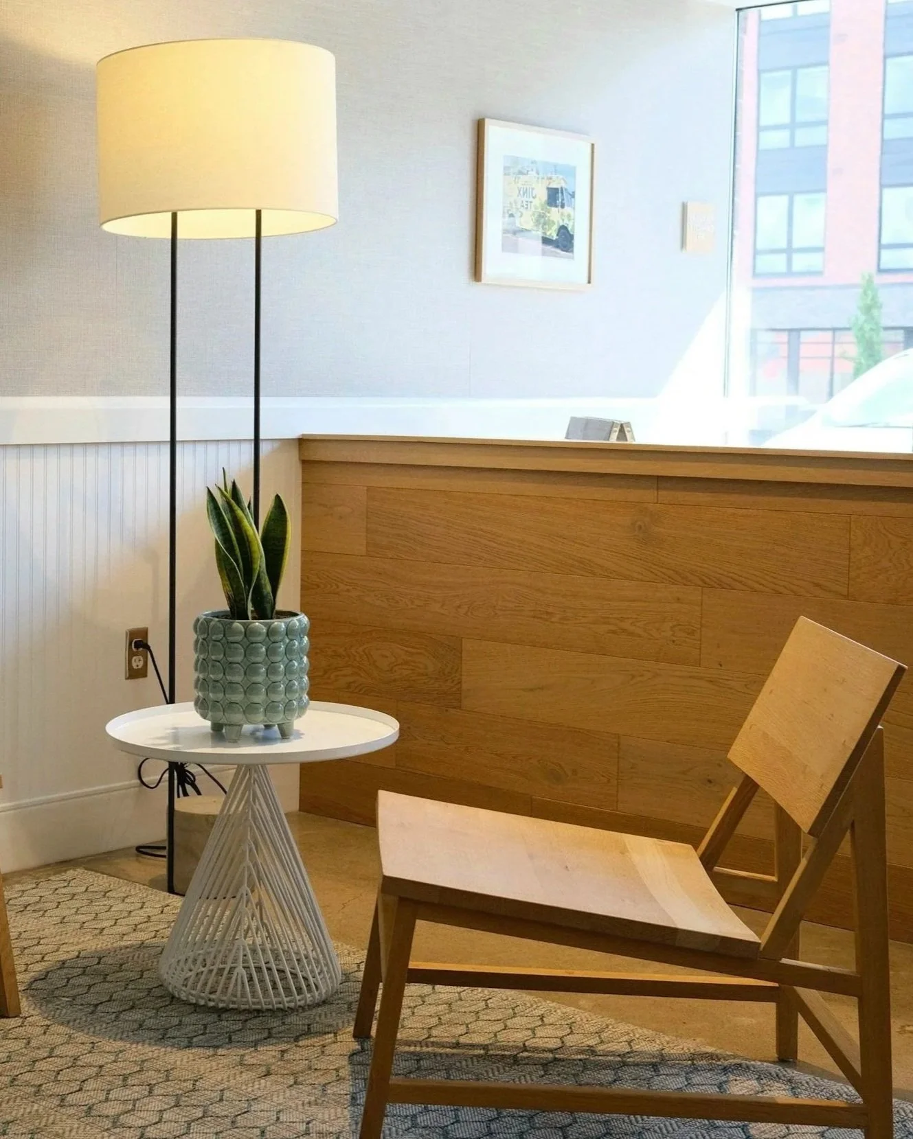 A cozy interior with a wooden chair, a small white side table with a decorative pot plant, a tall floor lamp, a wooden partition, a window showing a building outside, and a framed picture of a yellow vehicle on the wall.