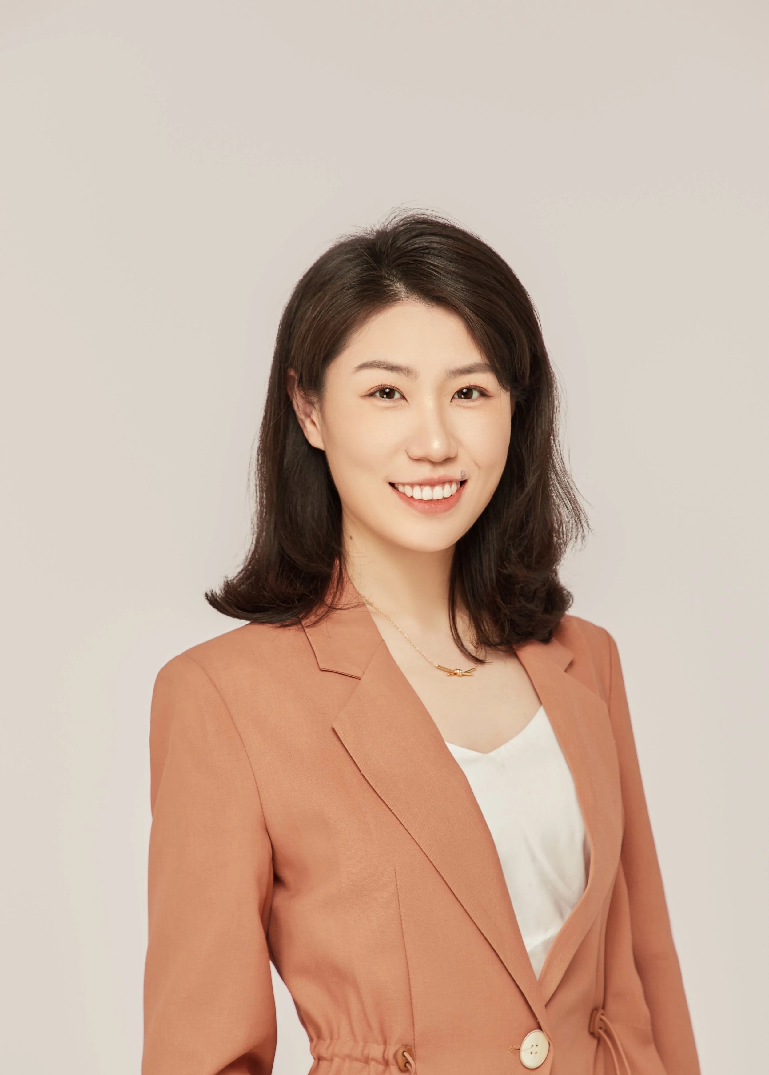 A woman with shoulder-length dark hair smiling, wearing a peach-colored blazer over a white top, and a delicate necklace, standing against a plain light background.