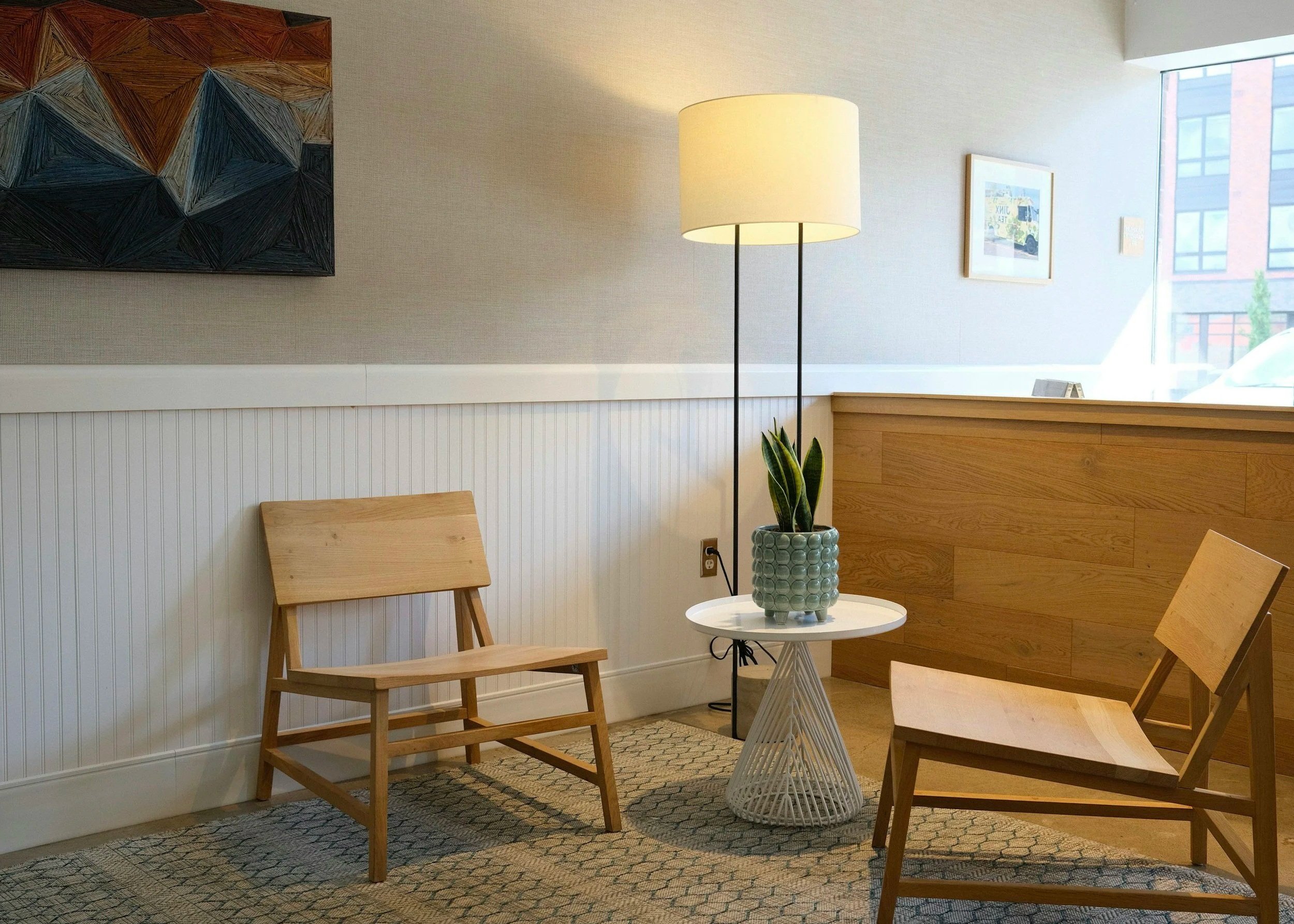 Interior of a modern waiting area featuring two wooden chairs, a standing lamp with a lampshade, a small round side table with a potted plant, a patterned carpet, and framed artwork on the wall near large windows.