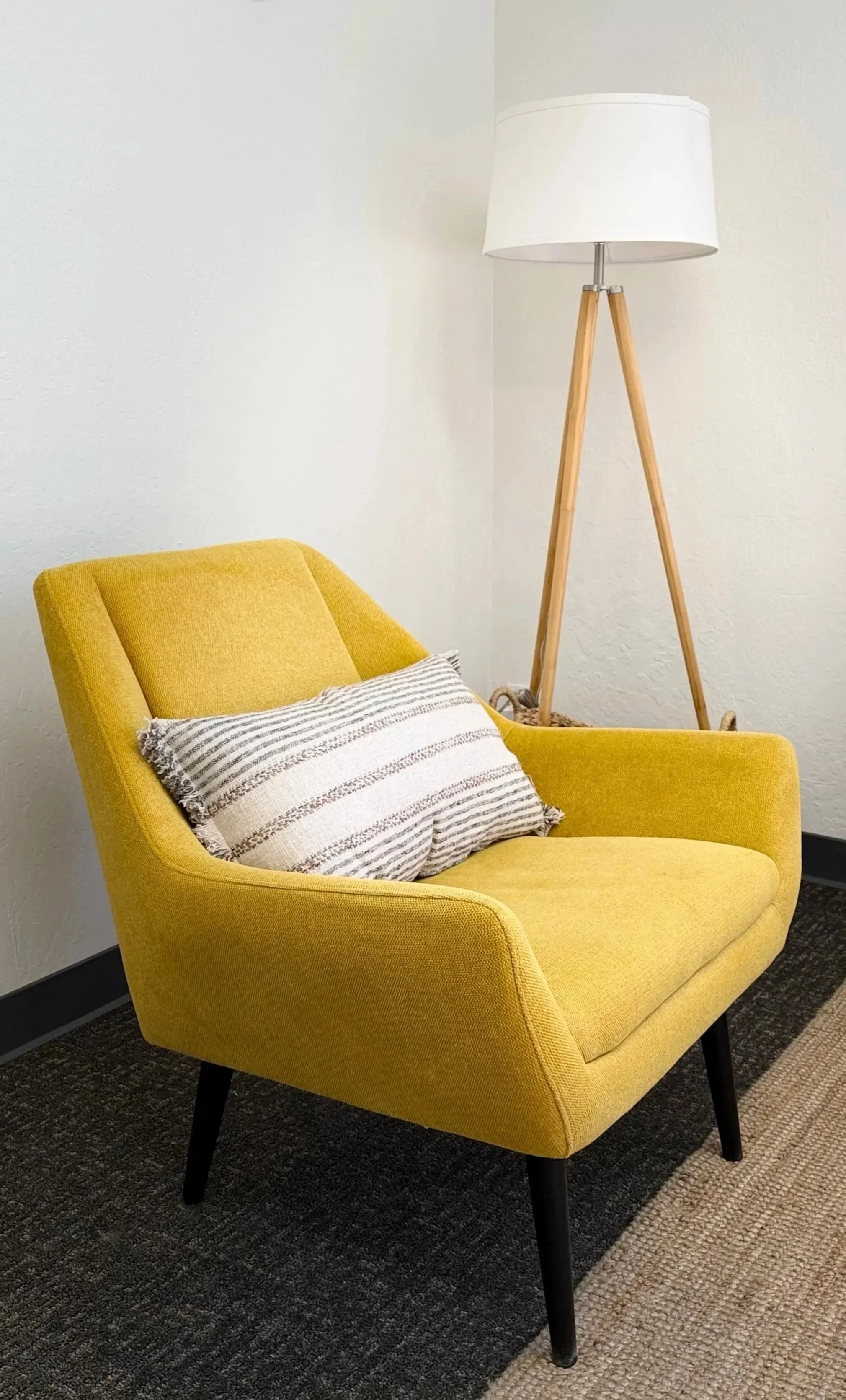 A yellow armchair with black legs, a striped pillow, and a tripod floor lamp with a white shade in a corner of a room with white walls and a multicolored carpet.