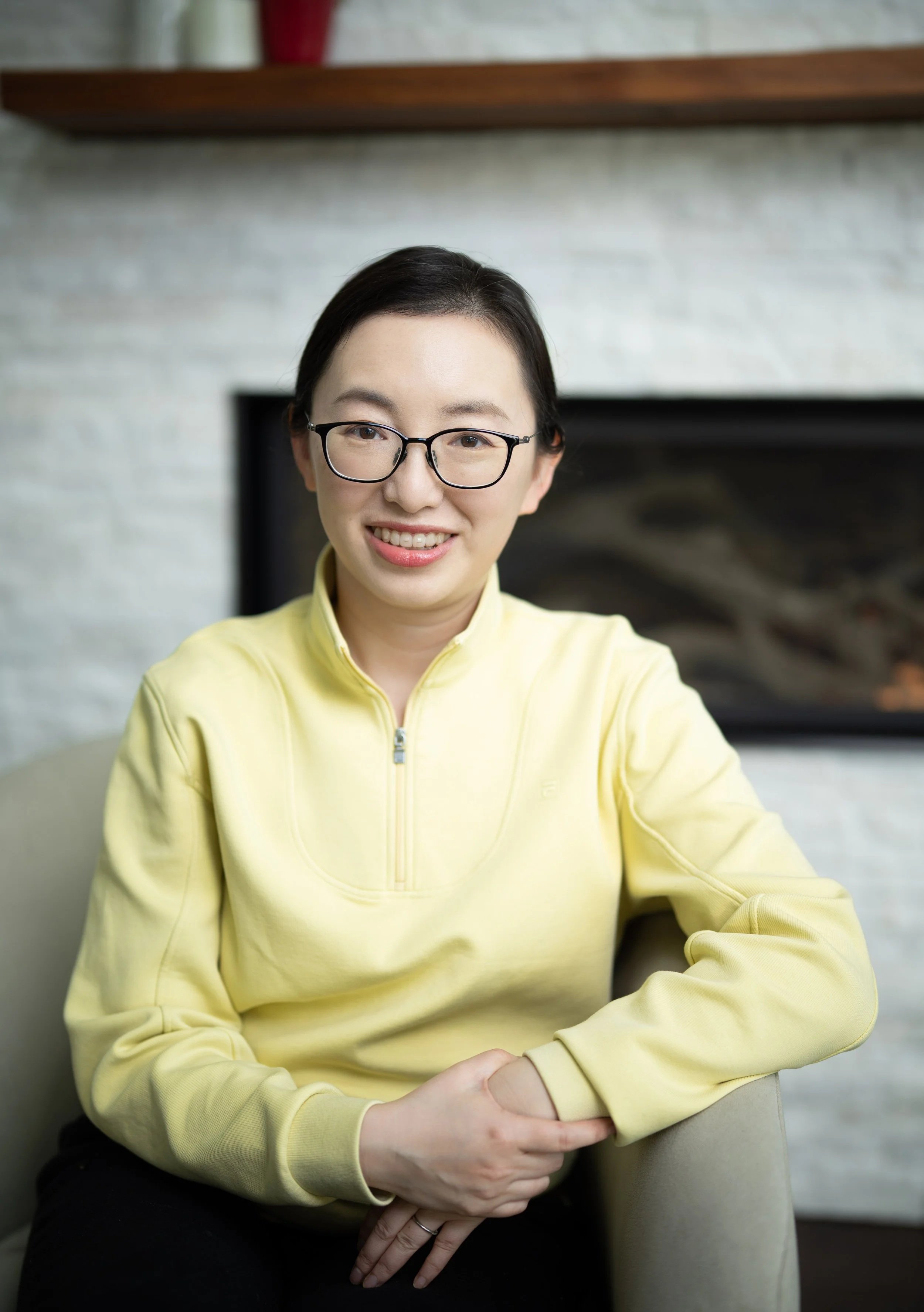 A woman wearing glasses and a yellow zip-up jacket sitting on a beige armchair, smiling, with a fireplace and a wooden shelf in the background.
