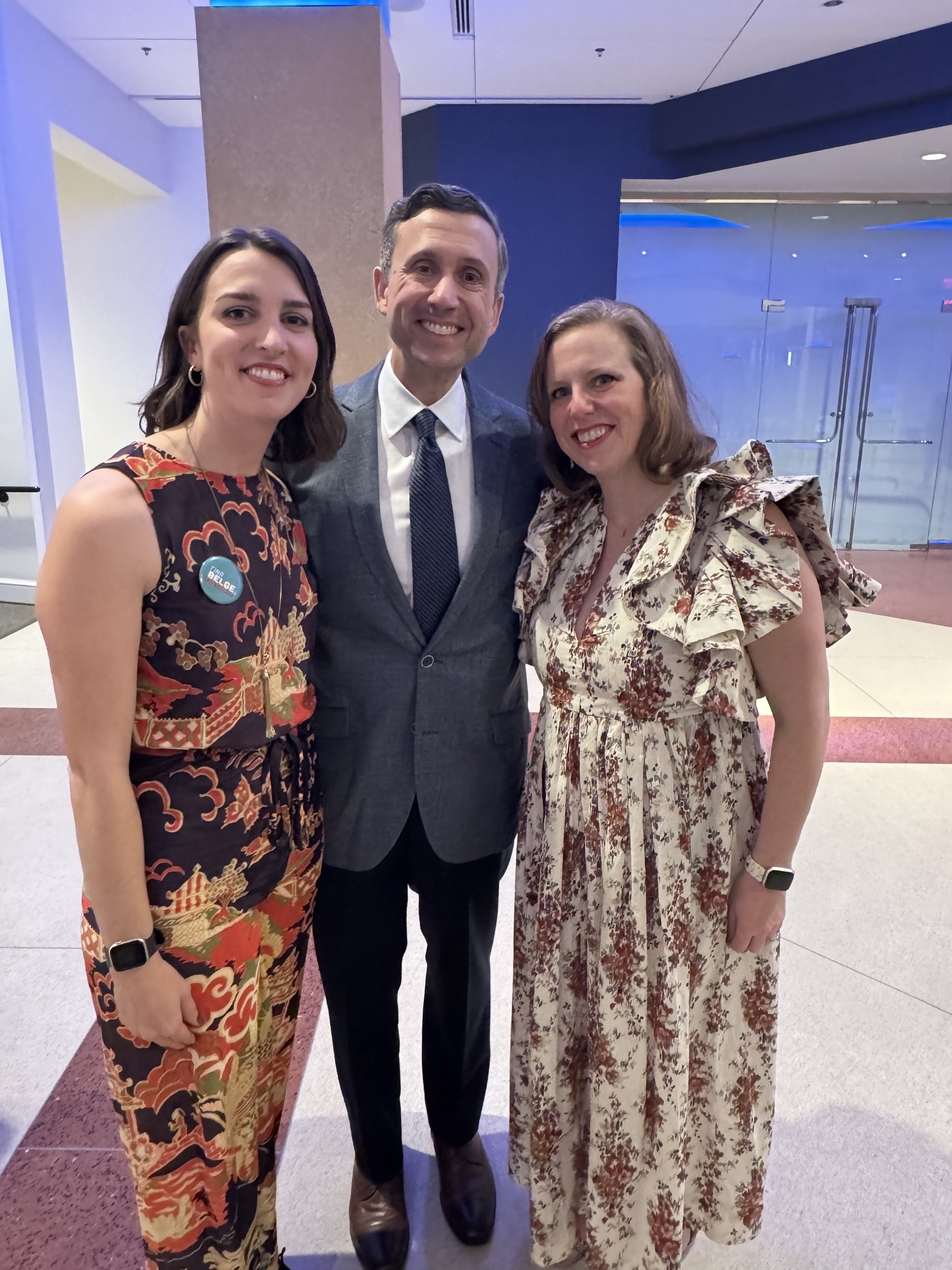 Three people posing together indoors; two women wearing patterned dresses and a man in a suit.