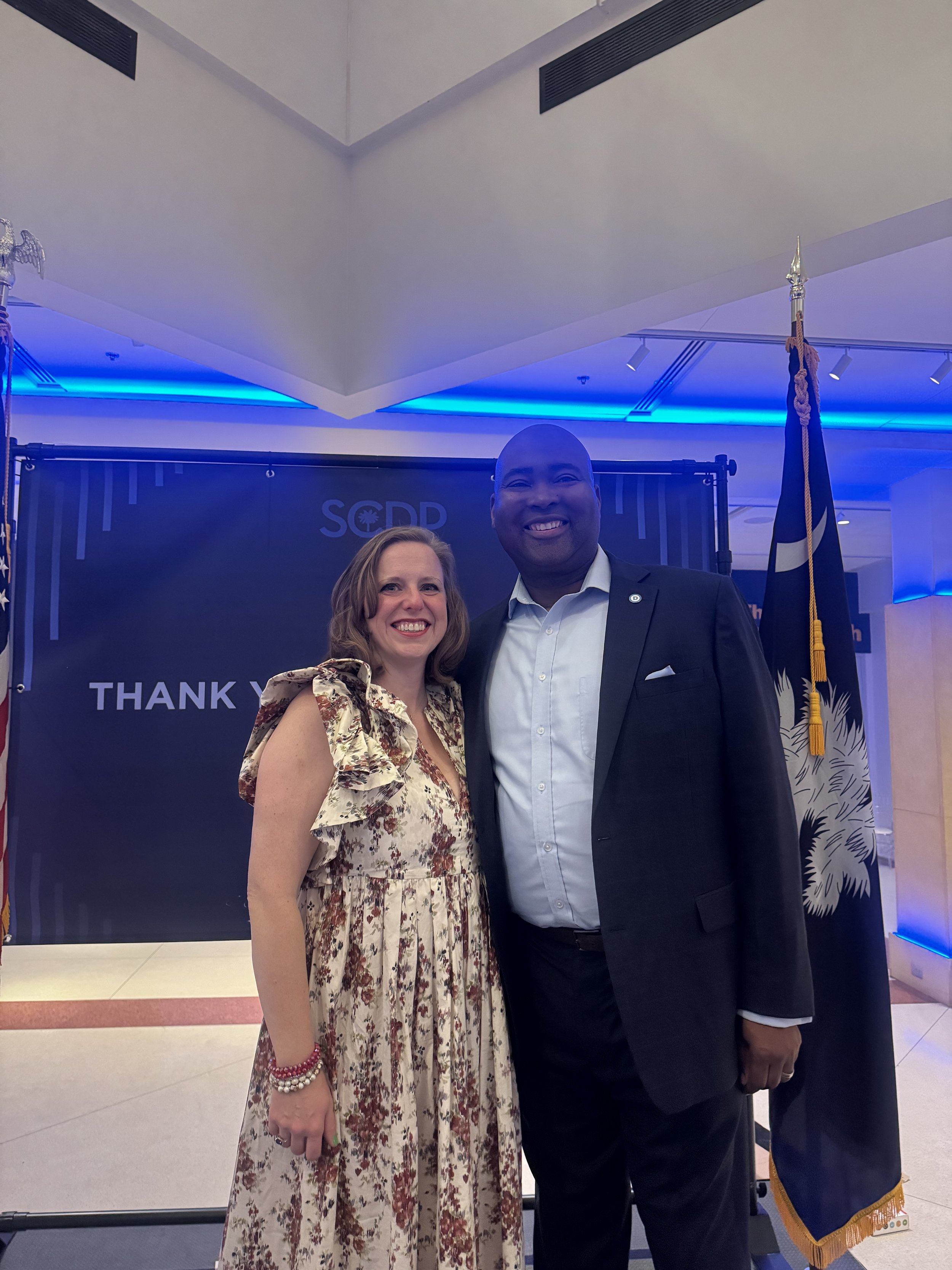 Two people standing together smiling in front of a backdrop with text "SGDD" and "THANK." The woman is wearing a floral dress, and the man is in a dark suit. American and South Carolina flags are visible.
