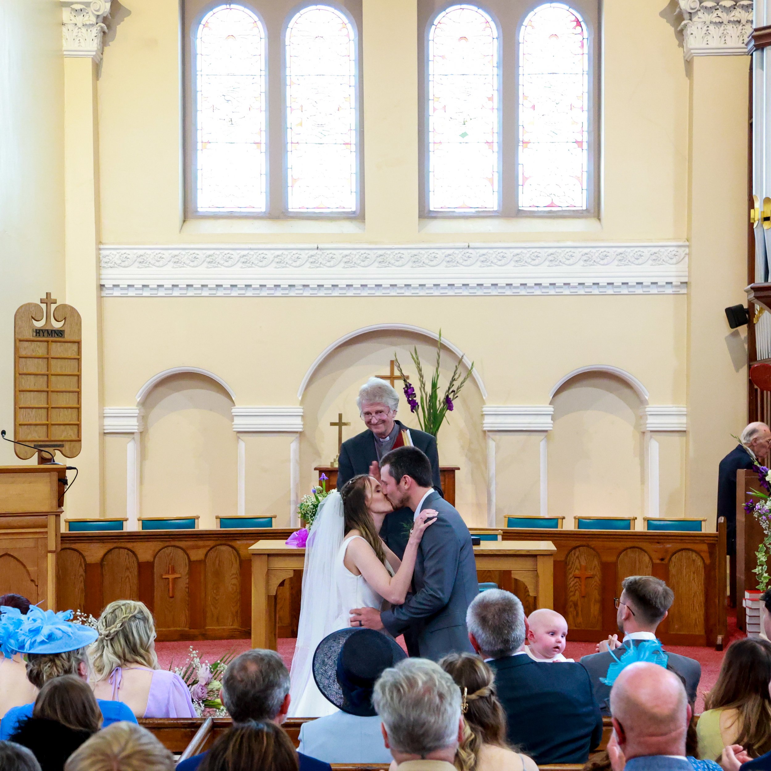 A bride and groom kissing during their wedding ceremony inside a church with stained glass windows, while a priest looks on smiling. Guests sit in pews watching the ceremony.