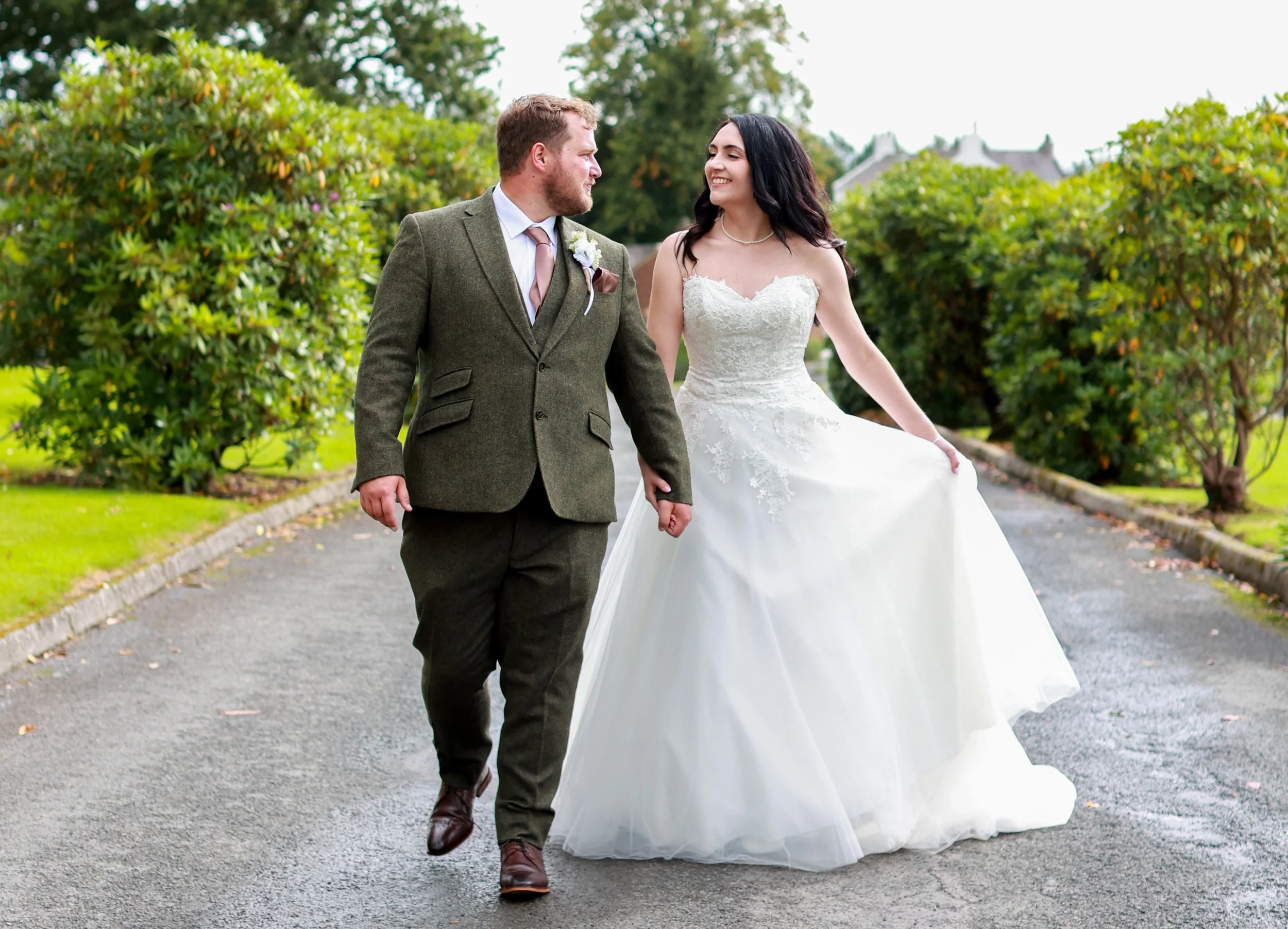 A bride and groom walking hand-in-hand outdoors on a cloudy day, with greenery and bushes in the background. The bride is wearing a white wedding gown, and the groom is dressed in a brown suit.