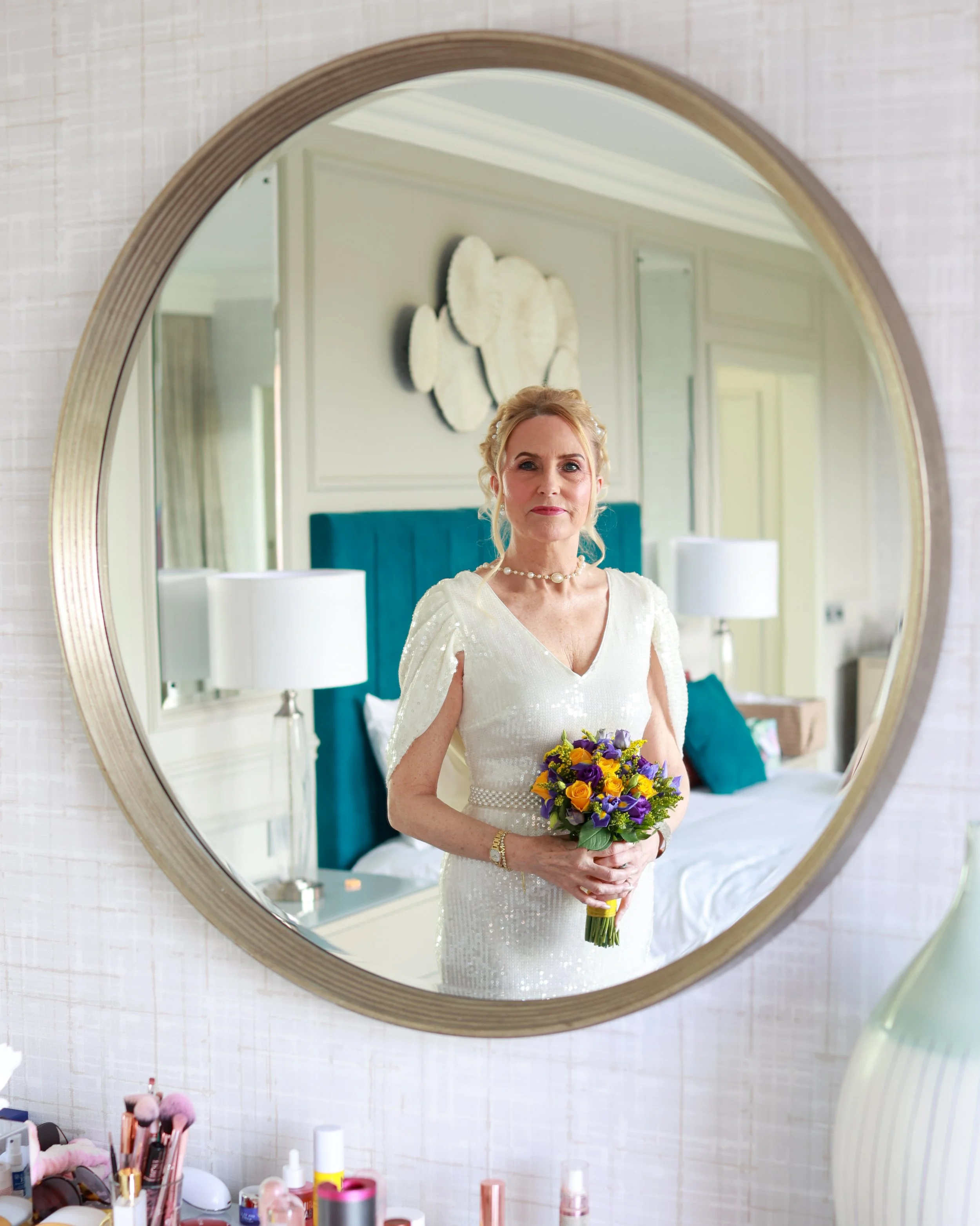 A woman in a white, sequined dress holding a bouquet of purple and yellow flowers, looking into a mirror in a bedroom with a teal headboard and lamps.