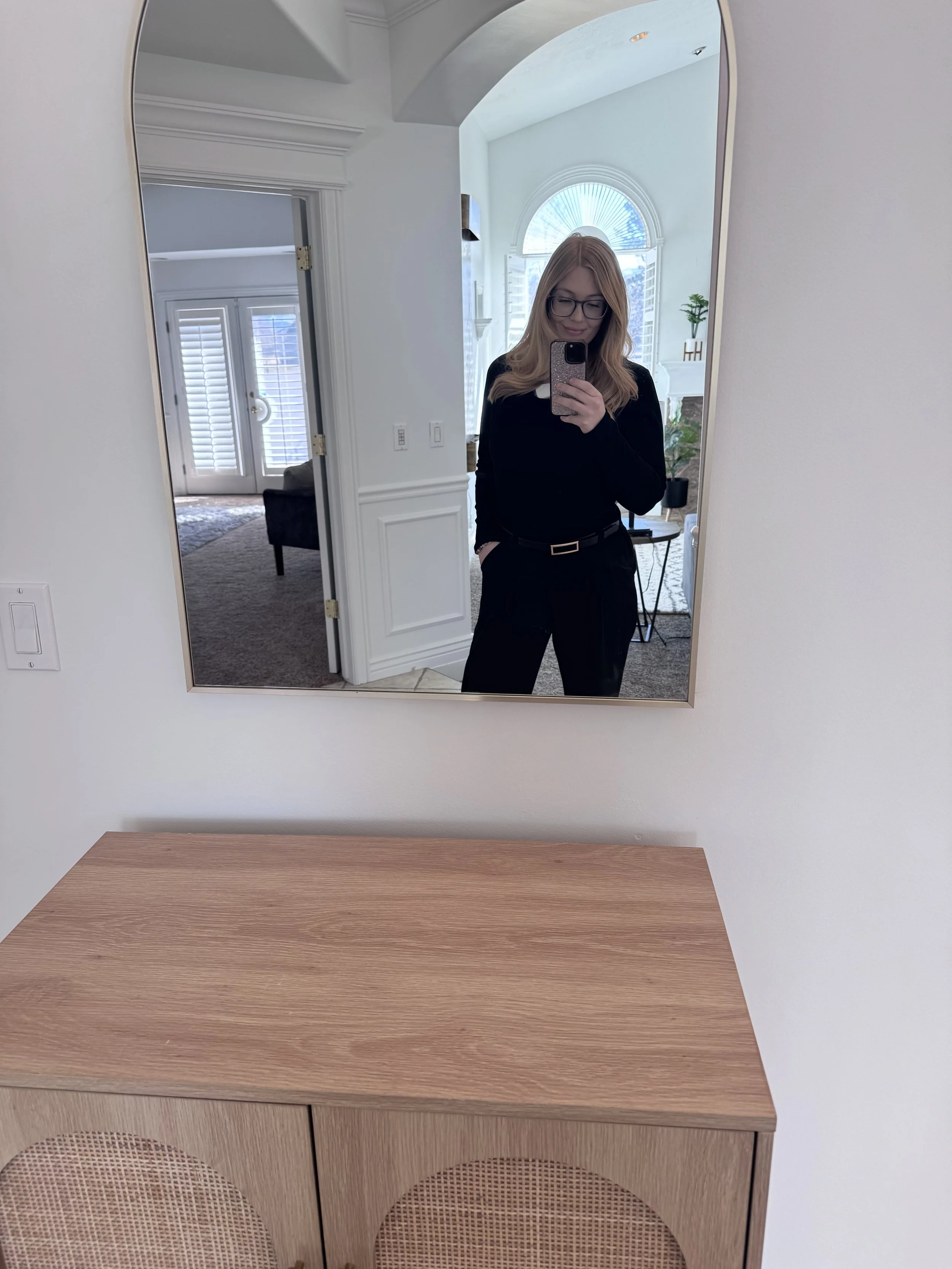 Person taking a mirror selfie in a room with a beige cabinet and arched mirror. The room has white walls and door frames, and a window with plantation shutters.