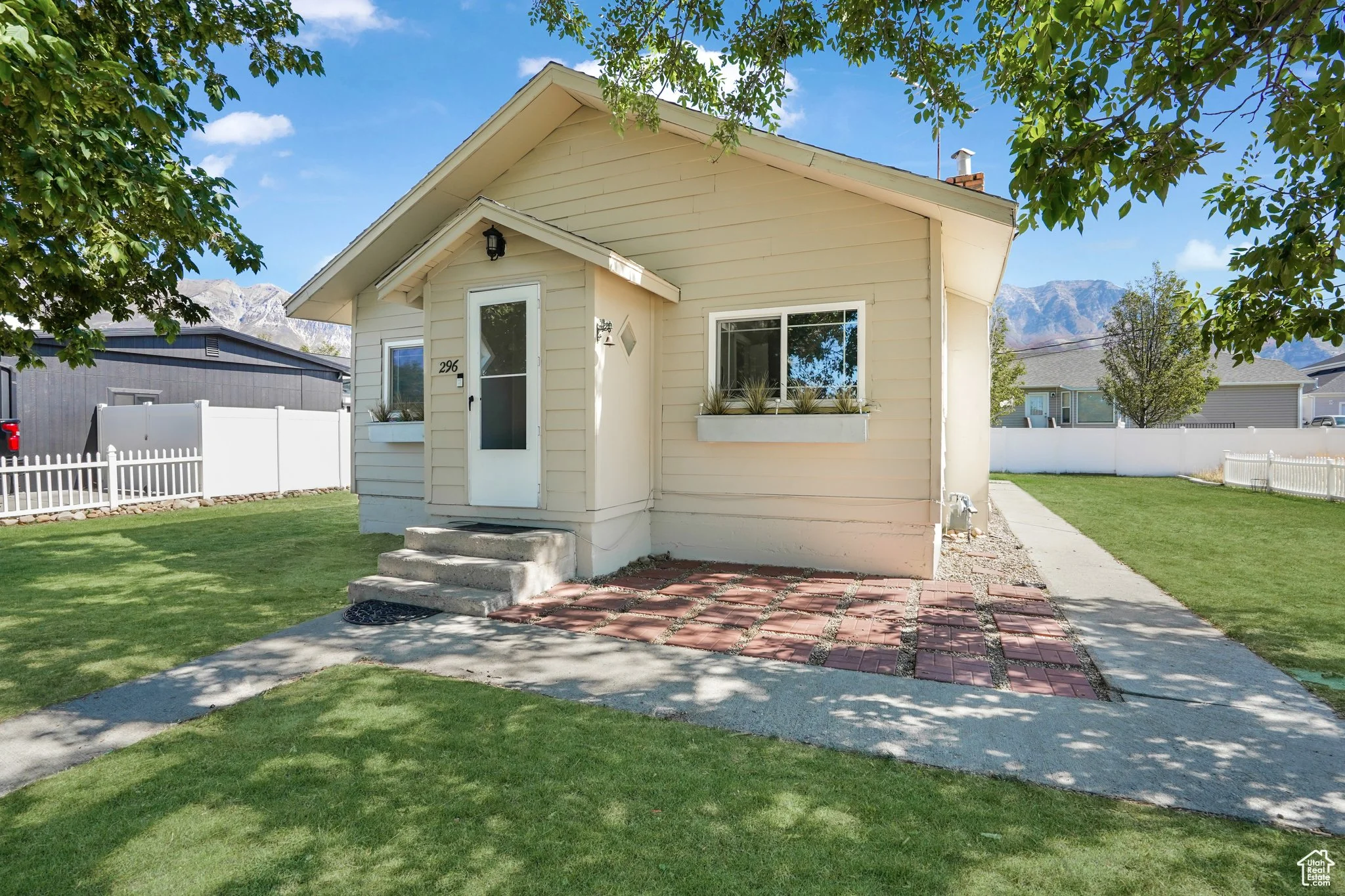 Single-story beige house with front porch and white door, surrounded by green lawn and trees, with mountains in the background.