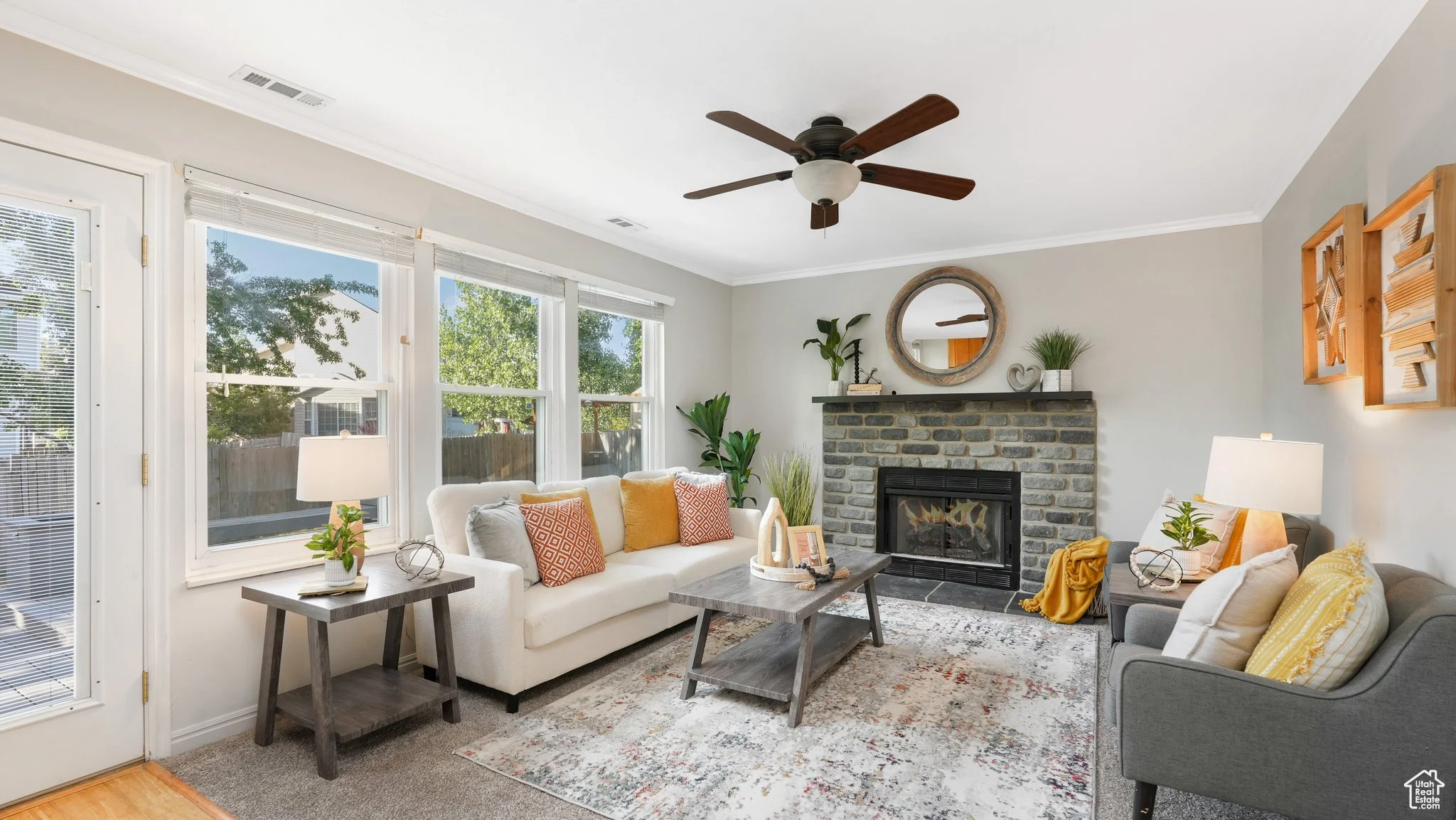 Modern living room with light gray walls, featuring a stone fireplace, ceiling fan, and large windows. The room has a white sofa with colorful cushions, a gray armchair, and wooden coffee and side tables. Decorative plants and a rug complete the cozy setting.