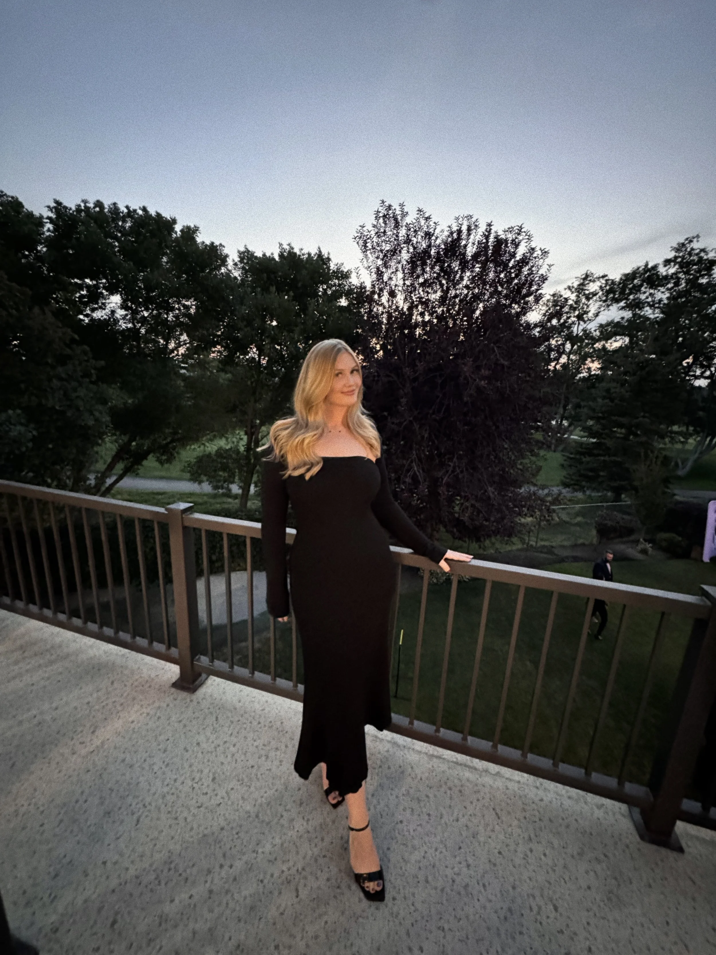 A woman in a black dress stands on a balcony with a dark sky and trees in the background.