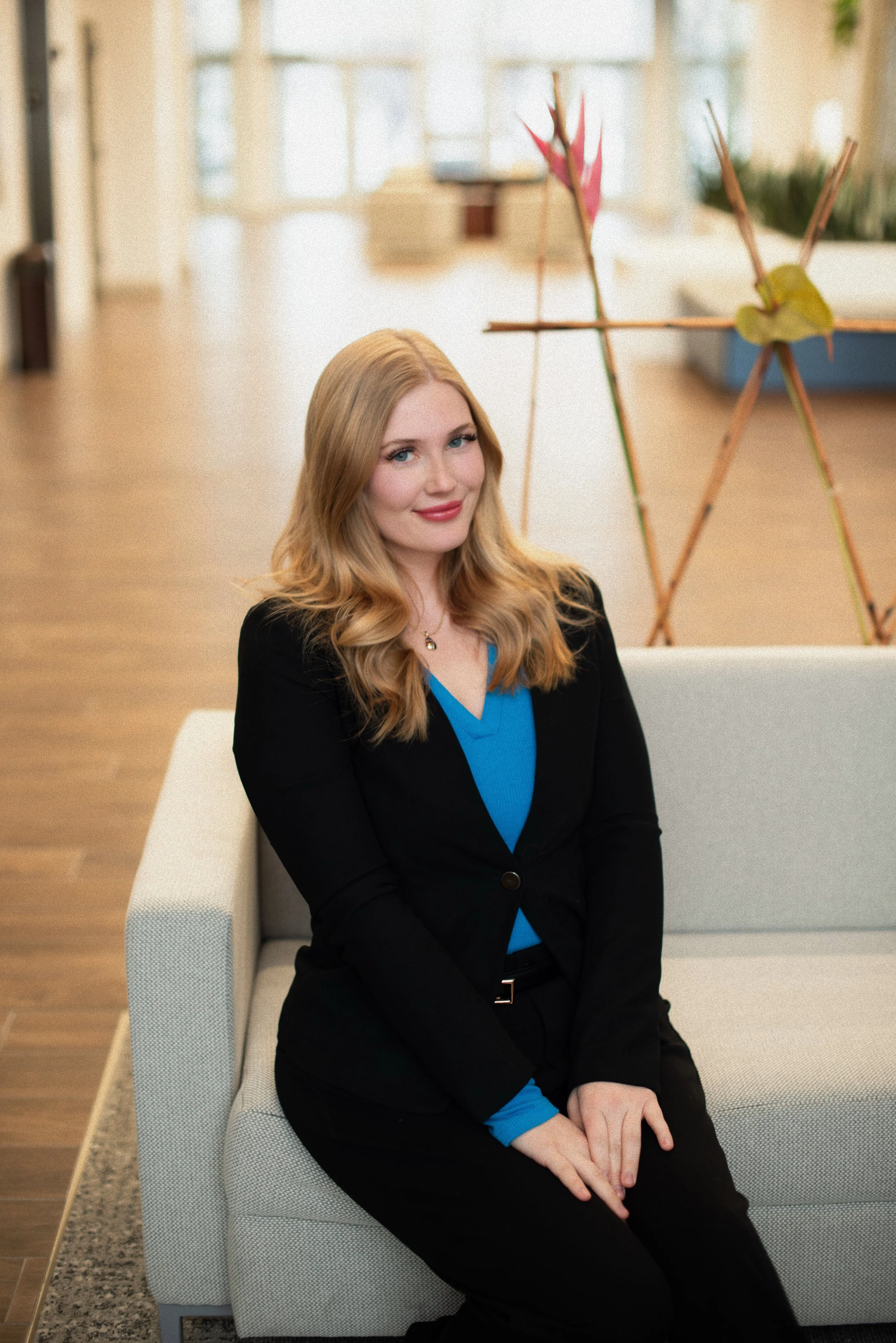 Businesswoman with long blonde hair seated on a light gray sofa in a modern office setting, wearing a black blazer and blue blouse.