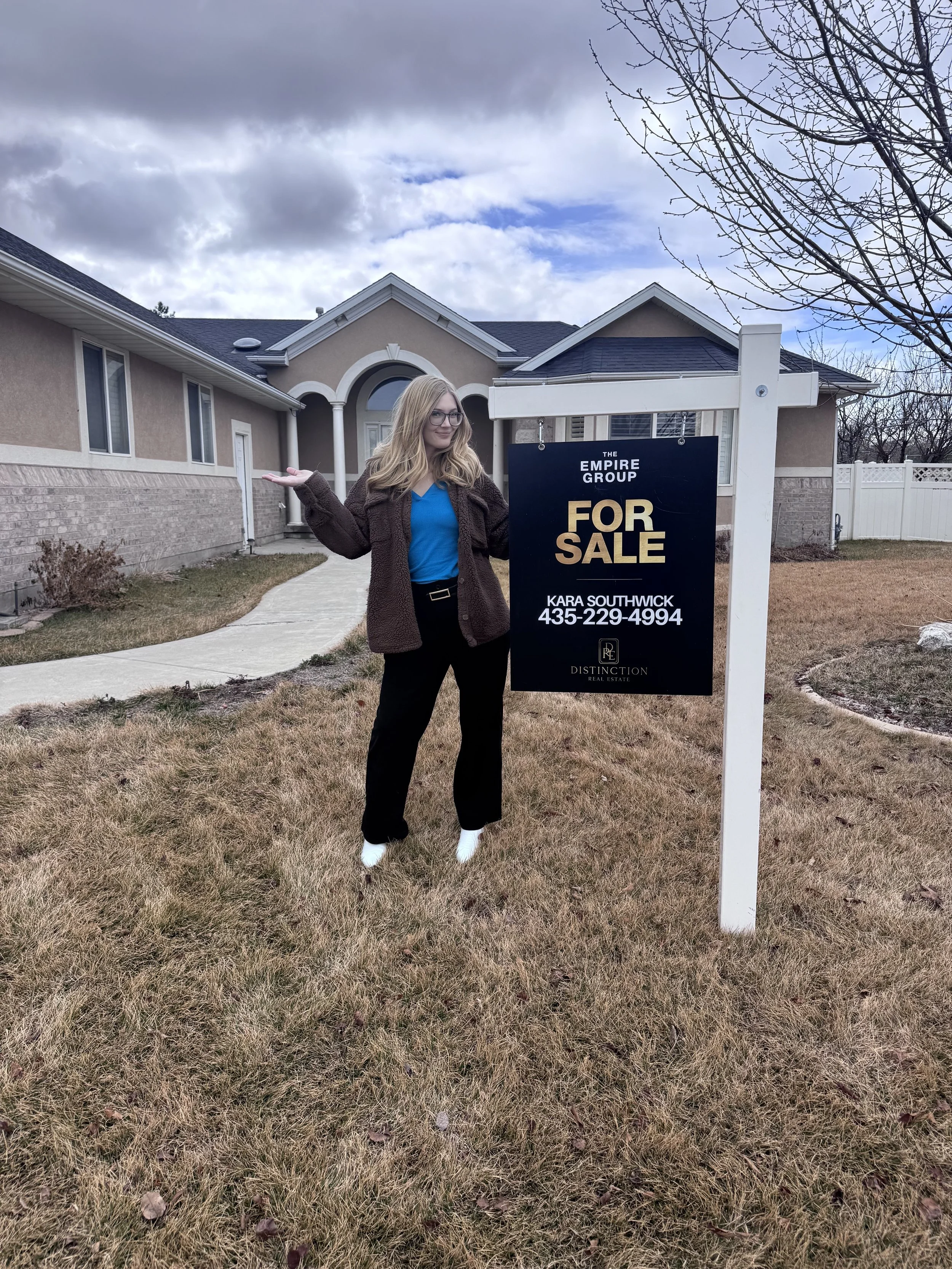 A woman stands in front of a house with a 'For Sale' sign by The Empire Group and Distinctive Real Estate, contact number visible. The lawn is brown, and the sky is overcast.
